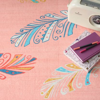 Pink patterned rug with blue and yellow feather motifs; white retro camera, purple notebook and pencils.