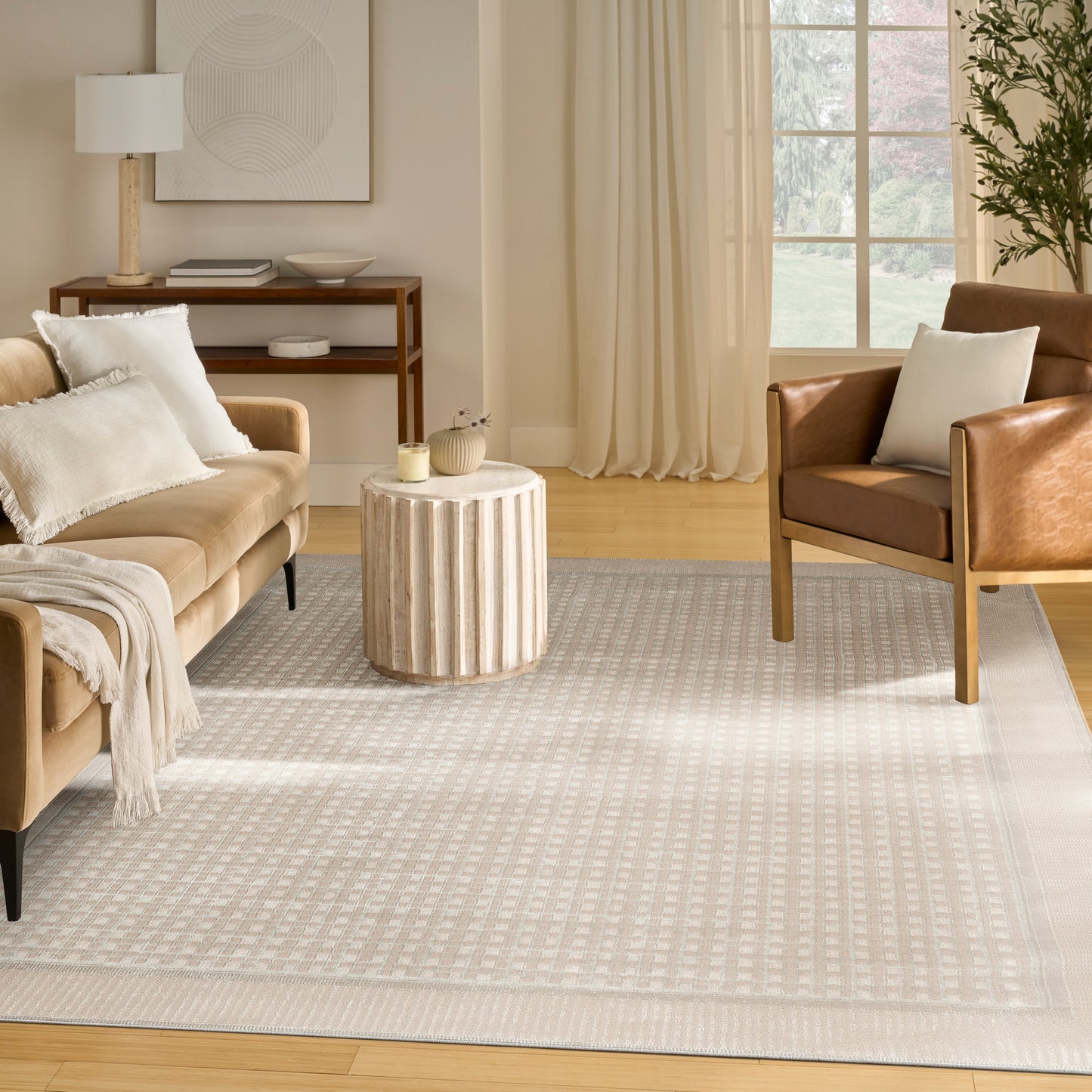 Neutral living room with beige sofa, round ribbed side table, light patterned rug, brown leather chair, and sunlit window.