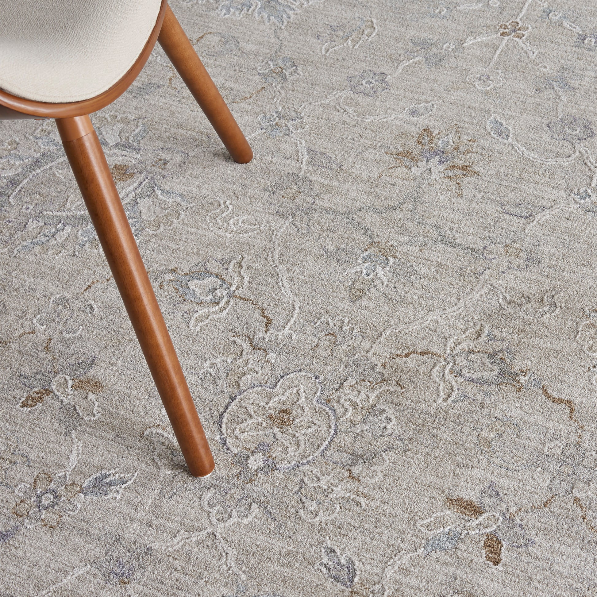 Brown wooden chair leg with beige upholstered seat on a beige floral rug with pale blue accents.