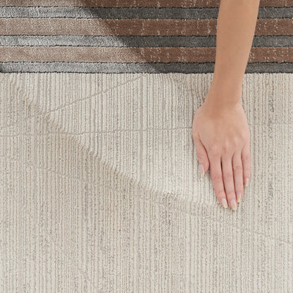 Beige textured rug with vertical stripes and brown border; hand with pale pink manicured nails resting on the rug.