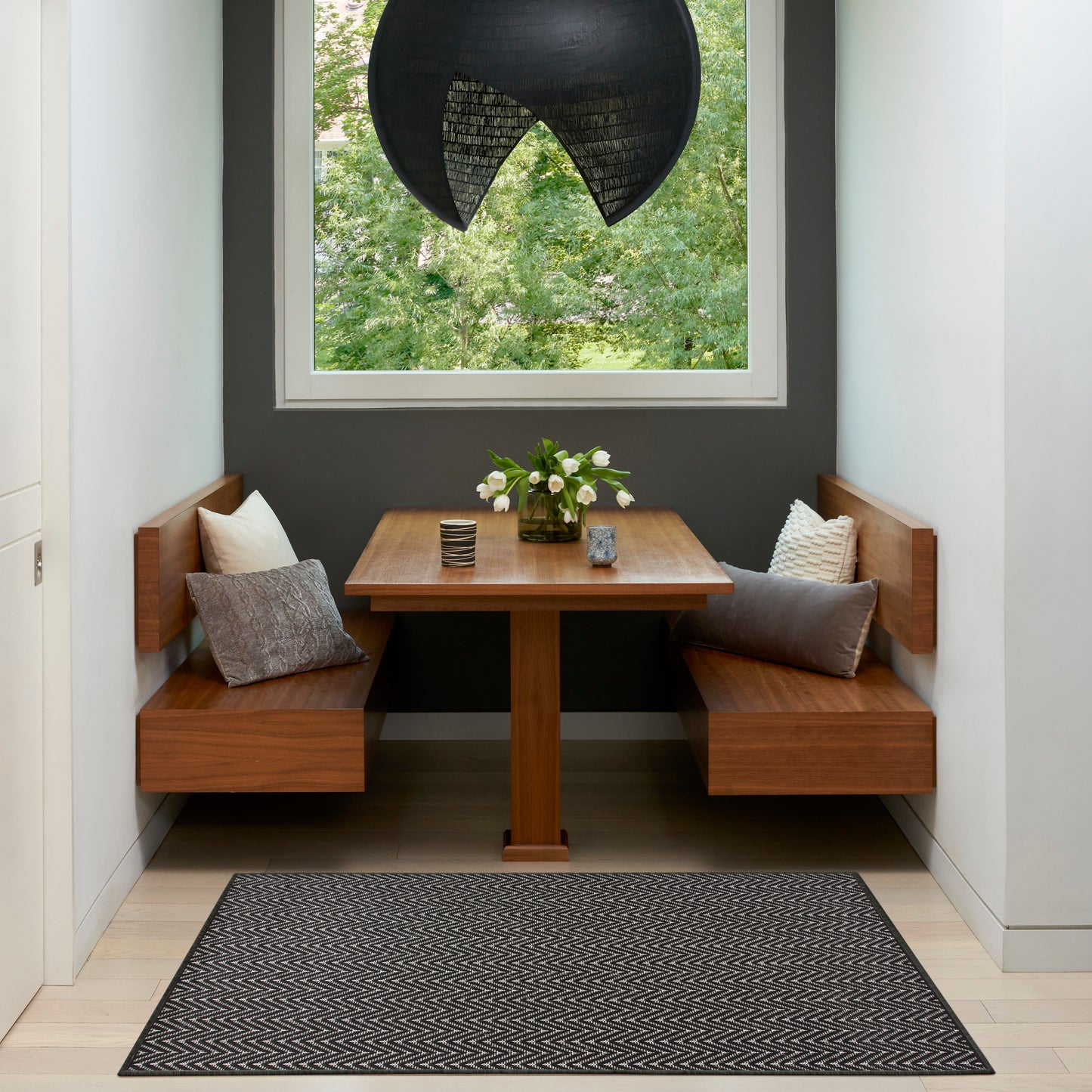 Chevron rug on a warm walnut dining nook with built-in benches, square table, gray cushions, flowers, dark wall, window, and a black pendant light.