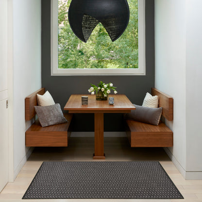 Chevron rug on a warm walnut dining nook with built-in benches, square table, gray cushions, flowers, dark wall, window, and a black pendant light.