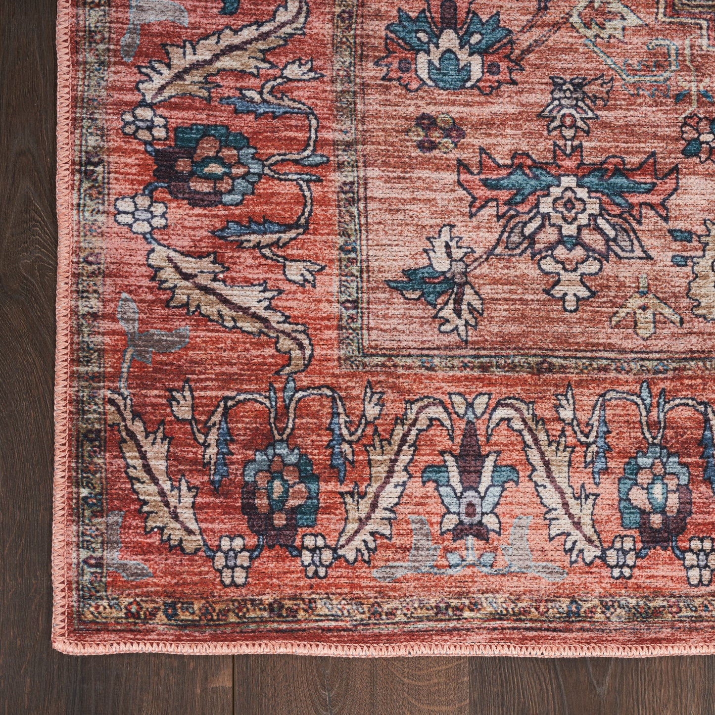 Oriental-style rug corner with red field, blue floral motifs, ivory border, pink fringe on dark wood floor.