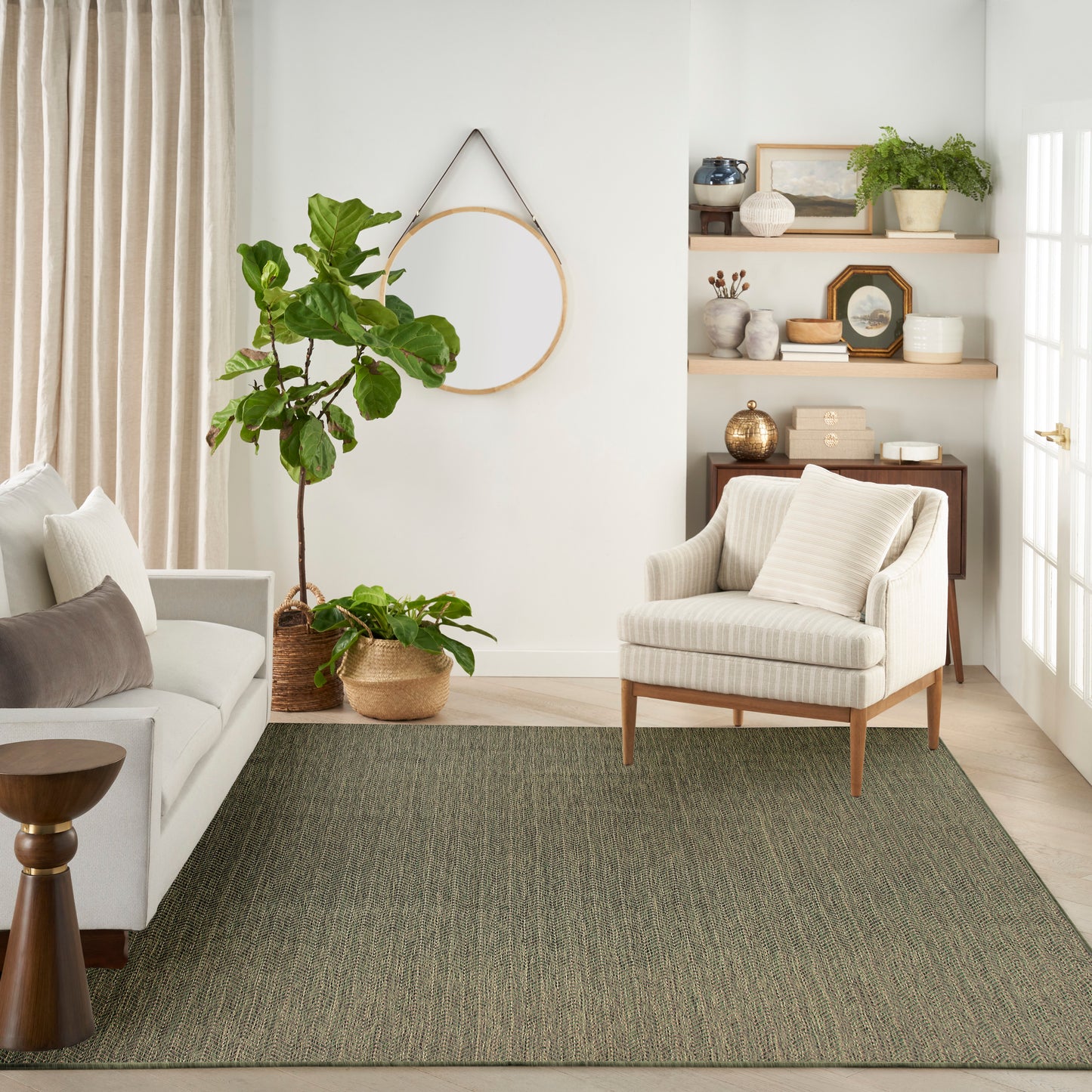 Cozy neutral living room with beige sofa and armchair, gray-green woven rug, potted plants, round wall hoop, wooden shelves.