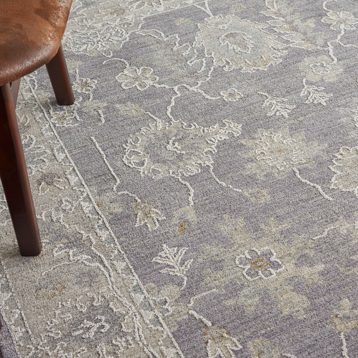 Gray-blue rug with cream floral damask pattern and border; brown leather chair leg visible.