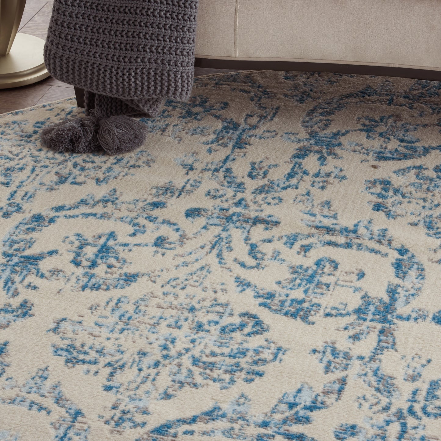 Beige rug with blue damask pattern; woven texture in a living room, foreground shows dark knit throw and fuzzy pom-pom slippers.