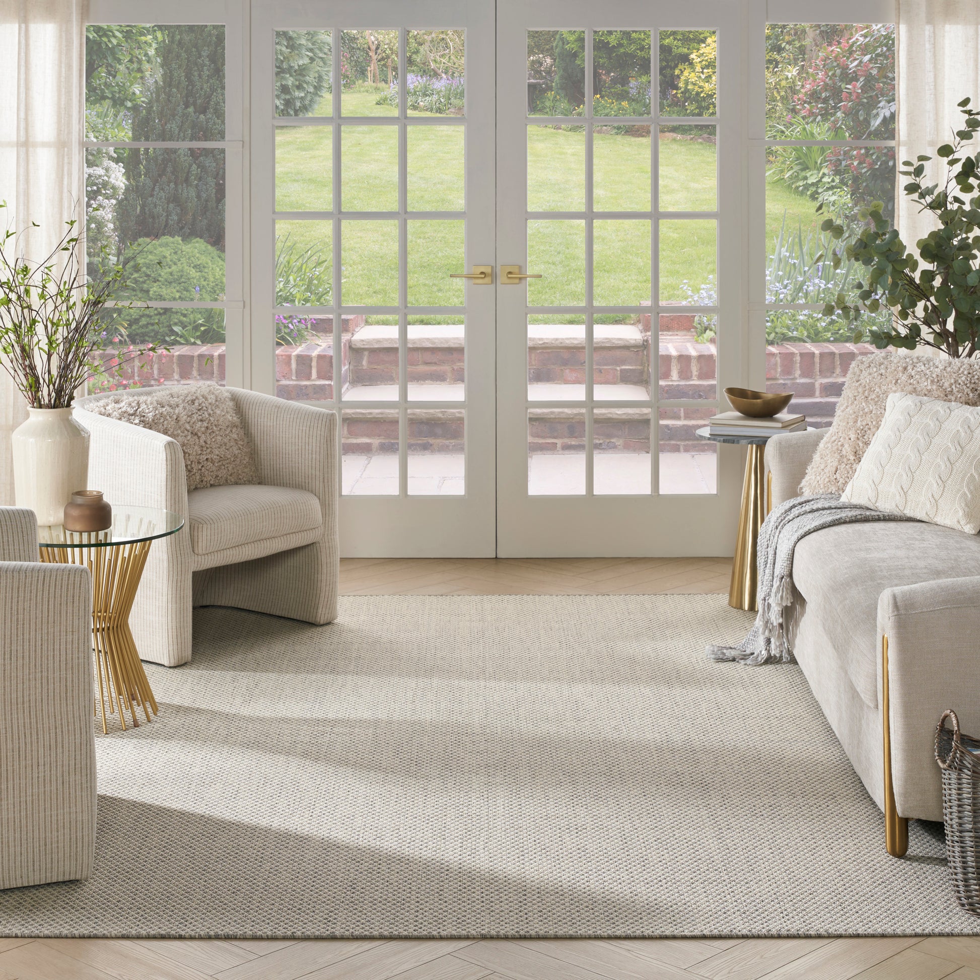 Bright neutral living room with cream sofa, beige armchairs, gold-legged side tables, light rug, and French doors to garden.