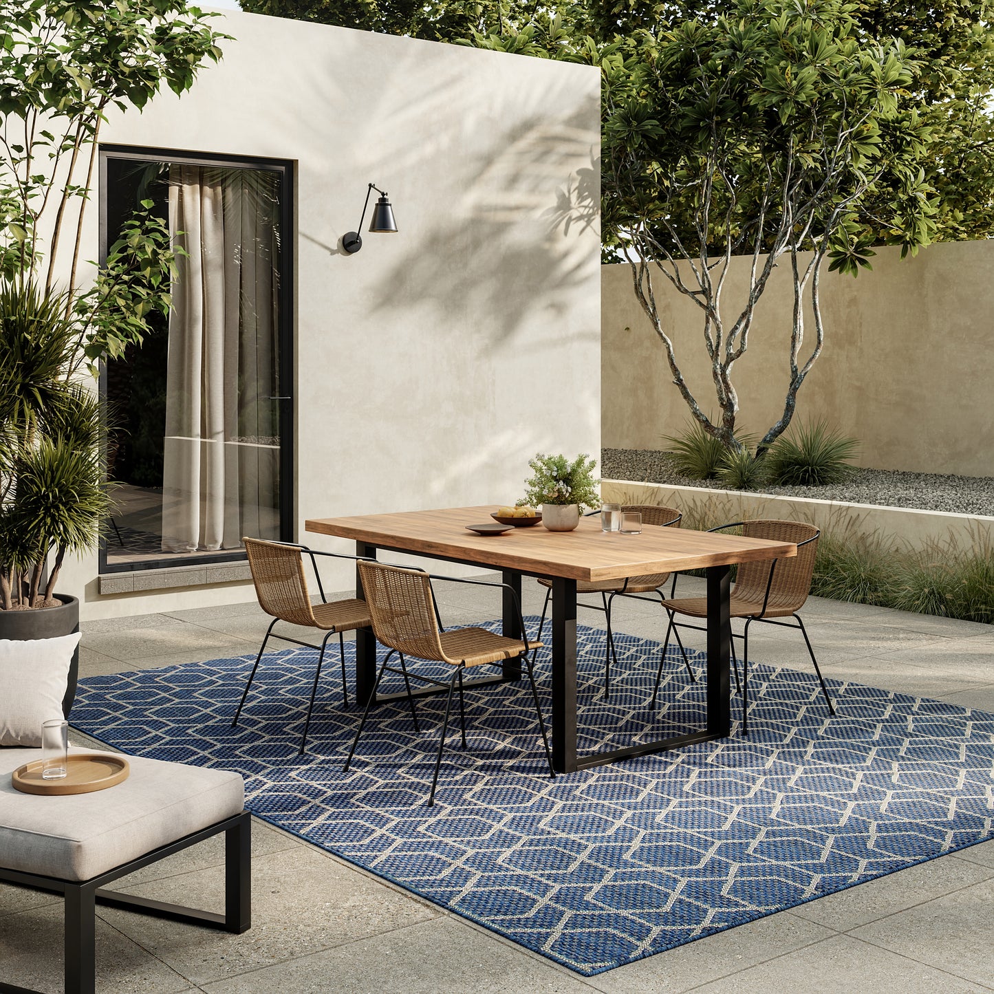 Outdoor dining area with wooden table and wicker chairs on a blue geometric rug, beige wall, lamp, and lush greenery.