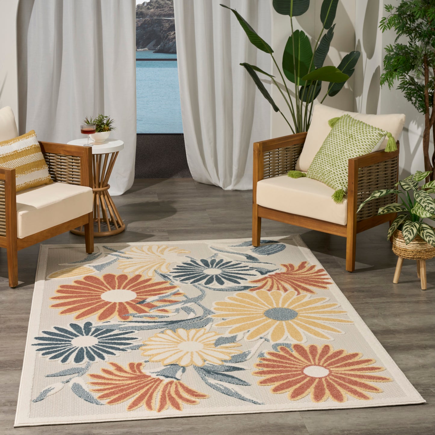 Cozy seating area with two wooden-framed armchairs, cream cushions, a colorful floral rug, potted plants, and sheer curtains by a water view.