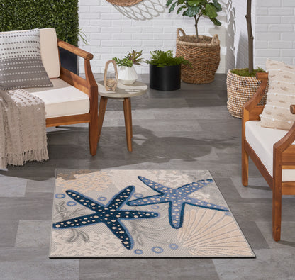 Coastal rug with blue starfish on beige sand background; wooden-framed chairs & neutral cushions, potted plants.