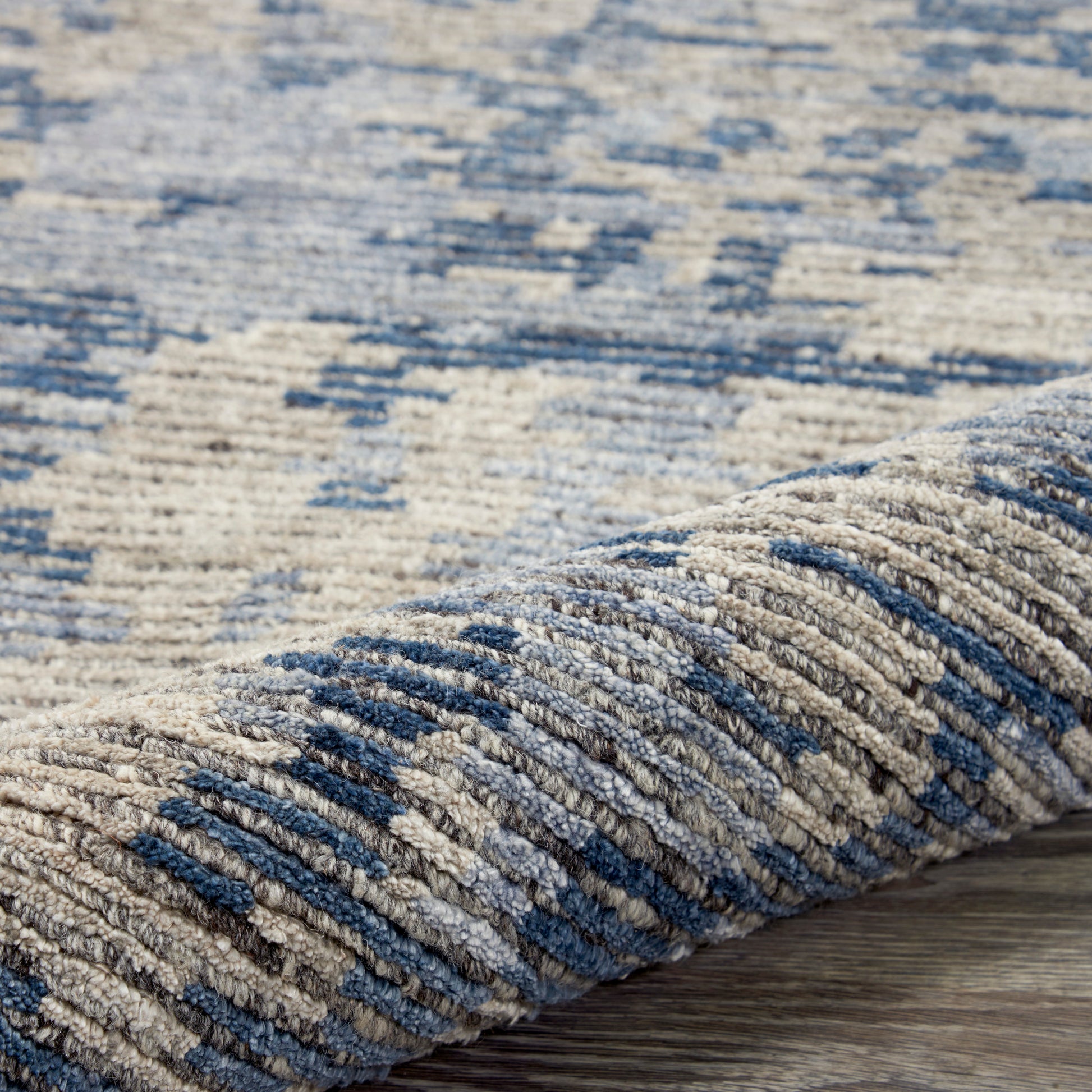 Close-up of a blue and beige geometric rug with a woven texture, rolled edge on a wood floor.