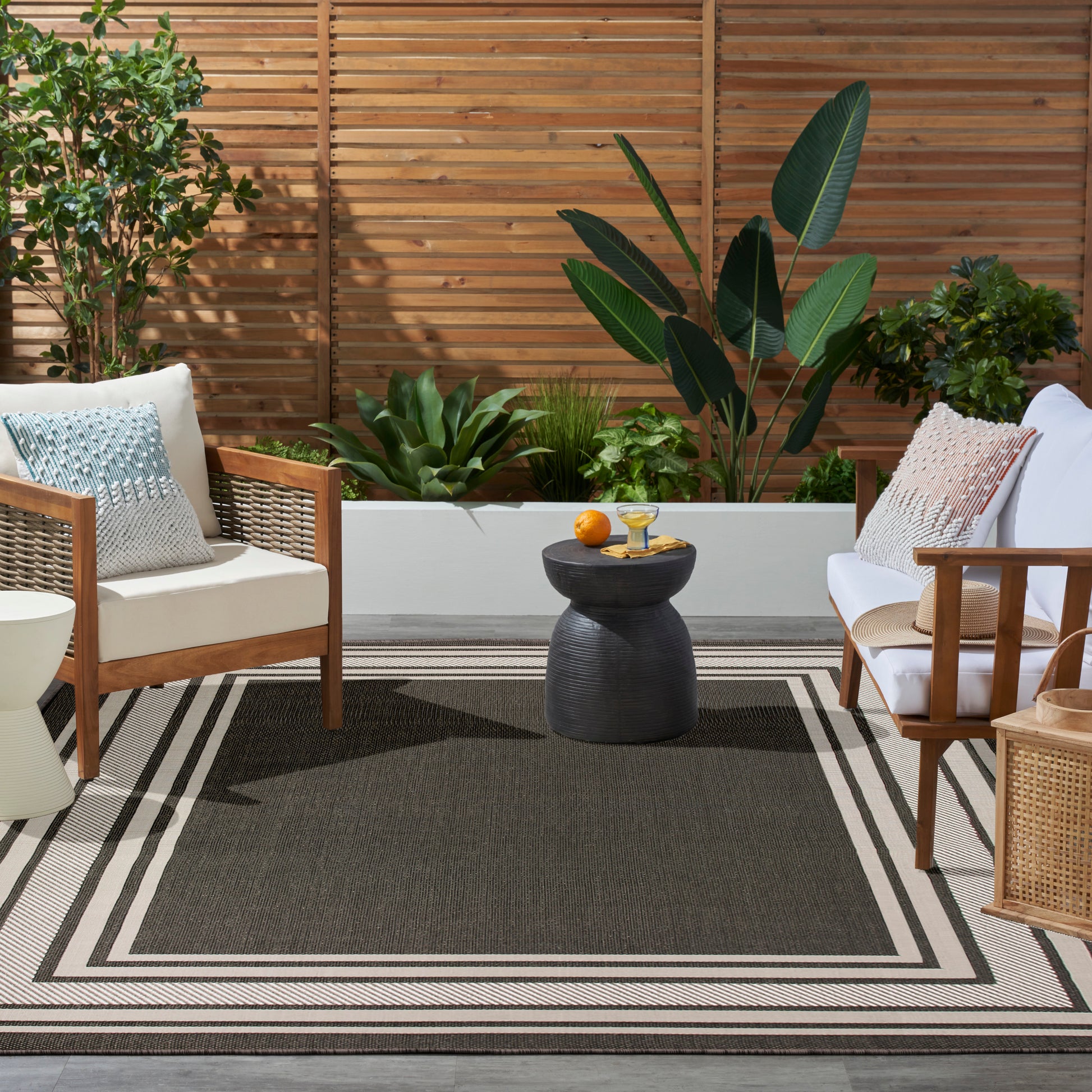 Outdoor patio with wooden-framed chairs, white cushions, black round side table, lush plants, and a striped outdoor rug.