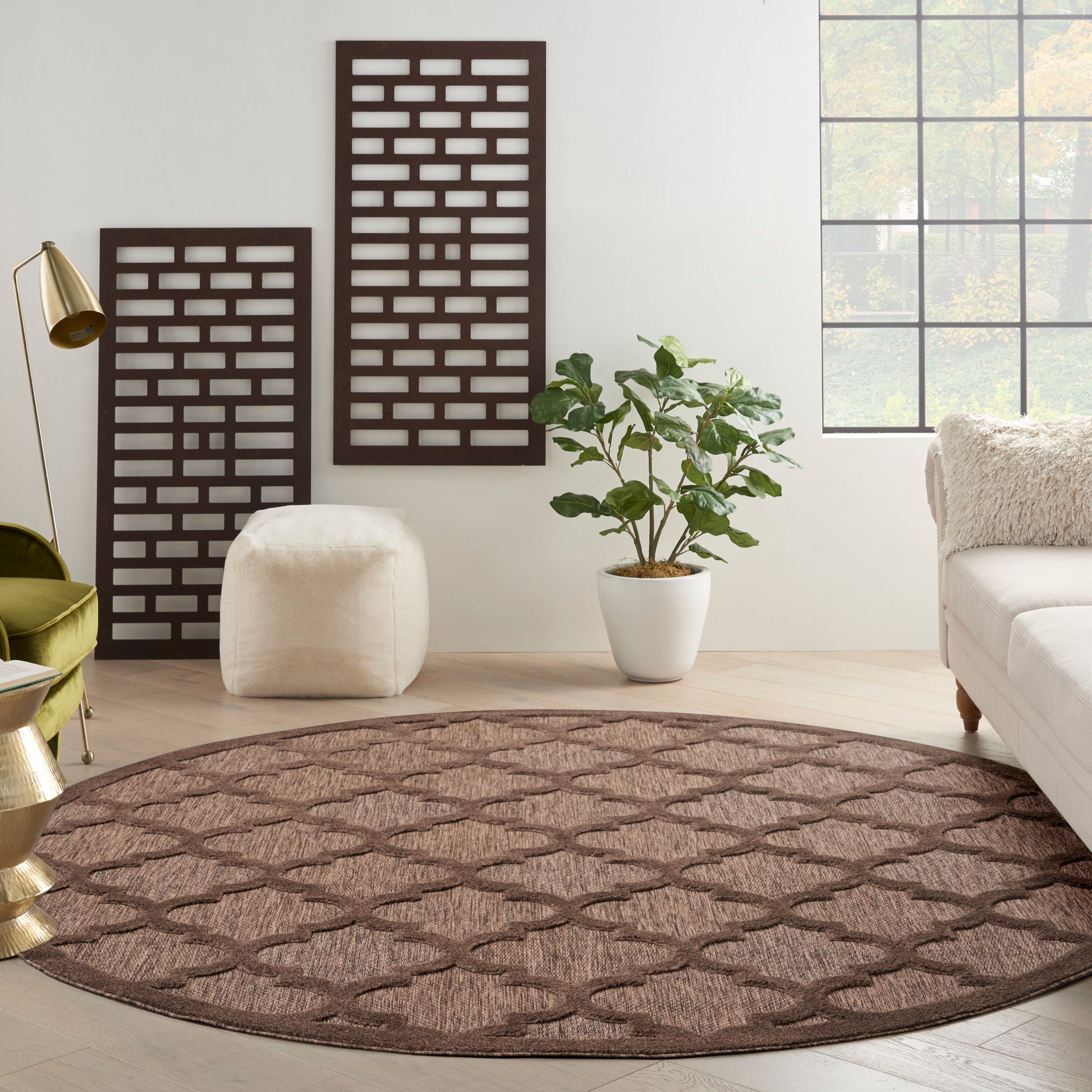 Round brown geometric rug in a bright living room with white walls, a potted plant, cream sofa, ottoman & gold lamp.