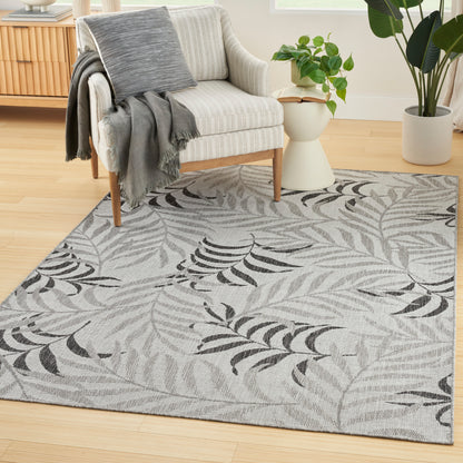 Light gray woven rug with black fern leaf design under a beige striped armchair; wooden legs, gray throw, and potted plants.
