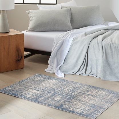 Rectangular blue-gray distressed area rug on light wood floor beside a white bed with gray duvet and pillows.