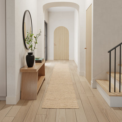 Bright hallway with light wood floors, round mirror, wooden console & vase, jute runner, arched door, stairs with black railing.