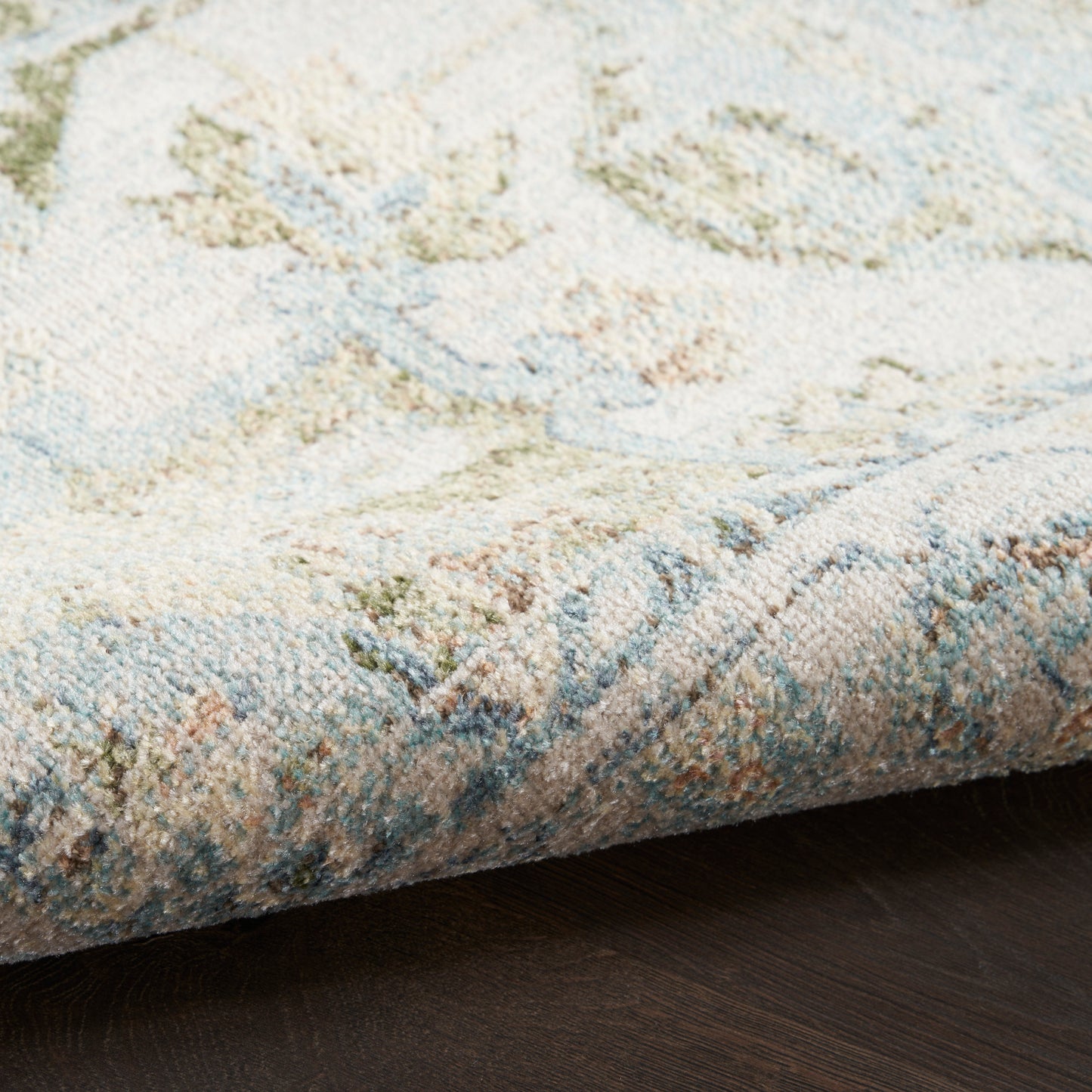 Pastel woven rug with cream base and blue, green, peach motifs; folded corner on dark wood floor.