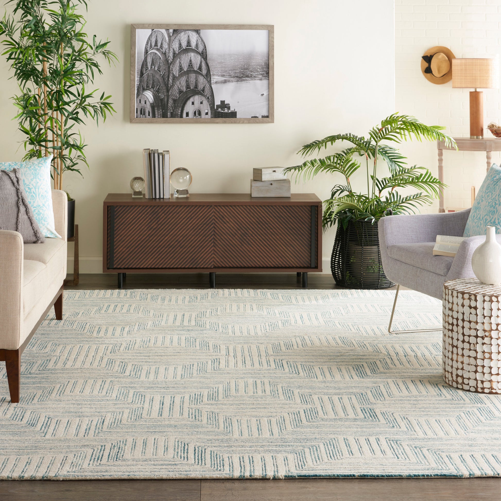 Living room with beige walls, chevron-front wood sideboard, black-and-white framed art, tall plants, pale sofa, blue geometric rug.