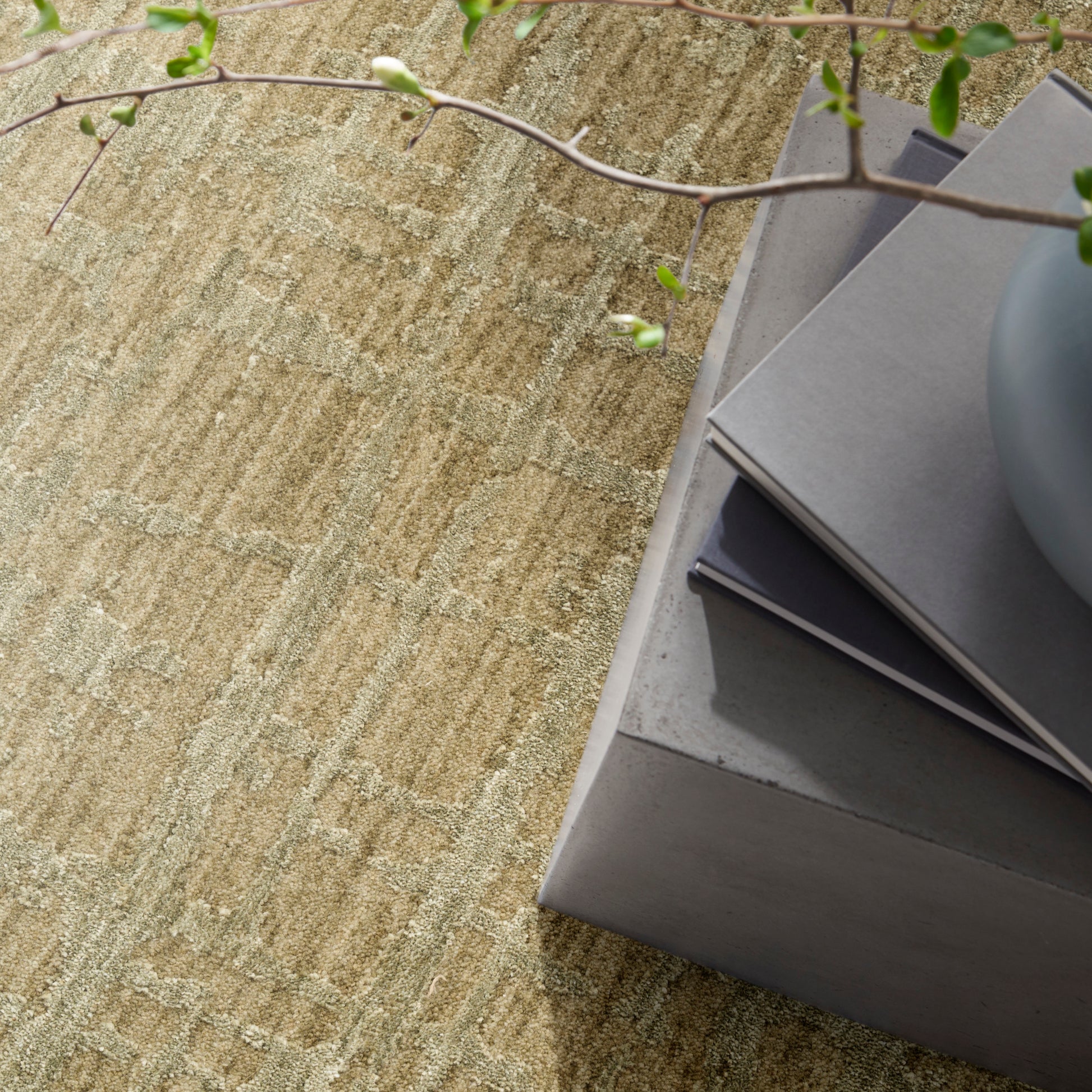 Beige textured rug, concrete pedestal with gray swatches, blue-gray vase, and trailing green plant branches.