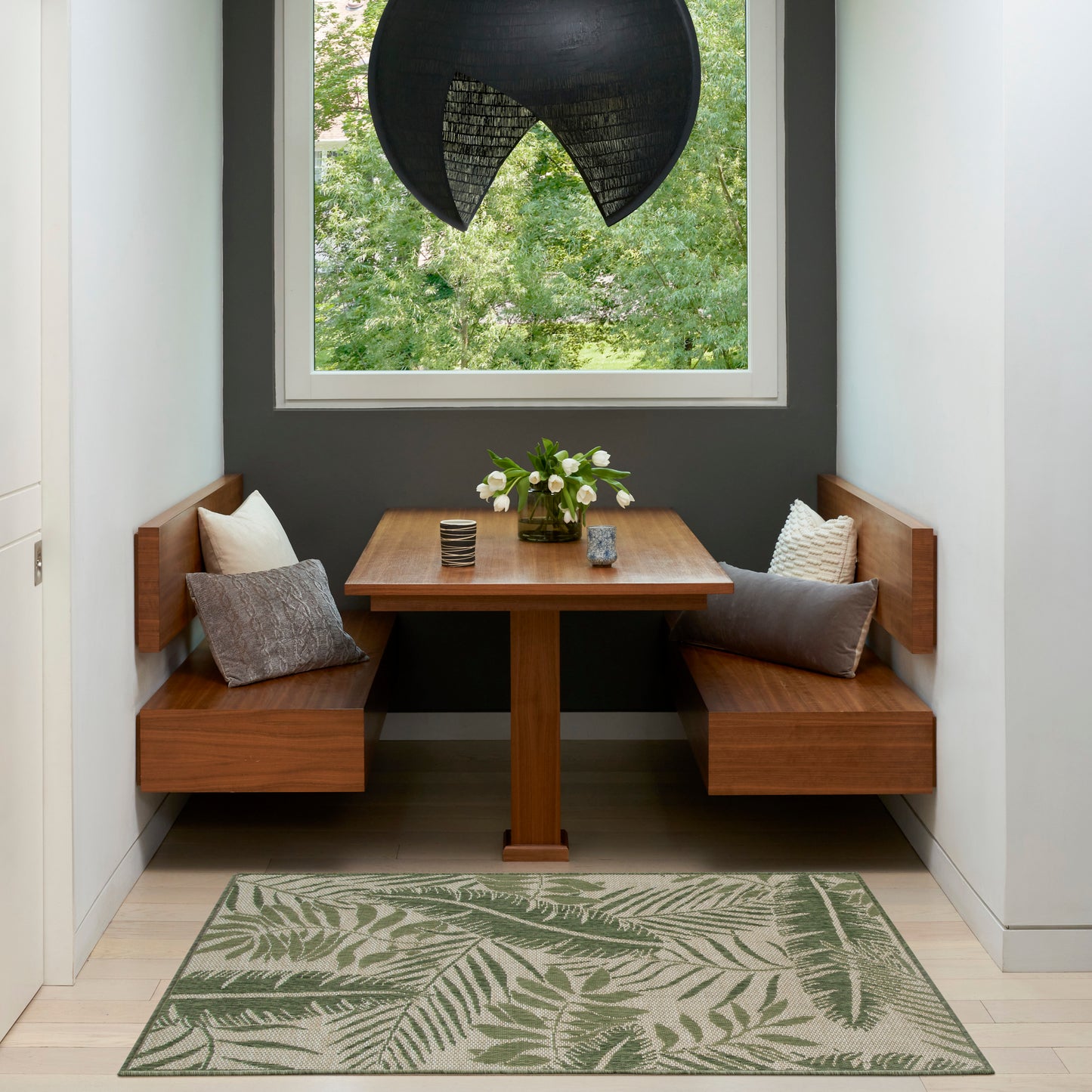 Wood dining nook with L-shaped benches, square table, gray cushions, leafy rug, dark wall and black woven pendant light.