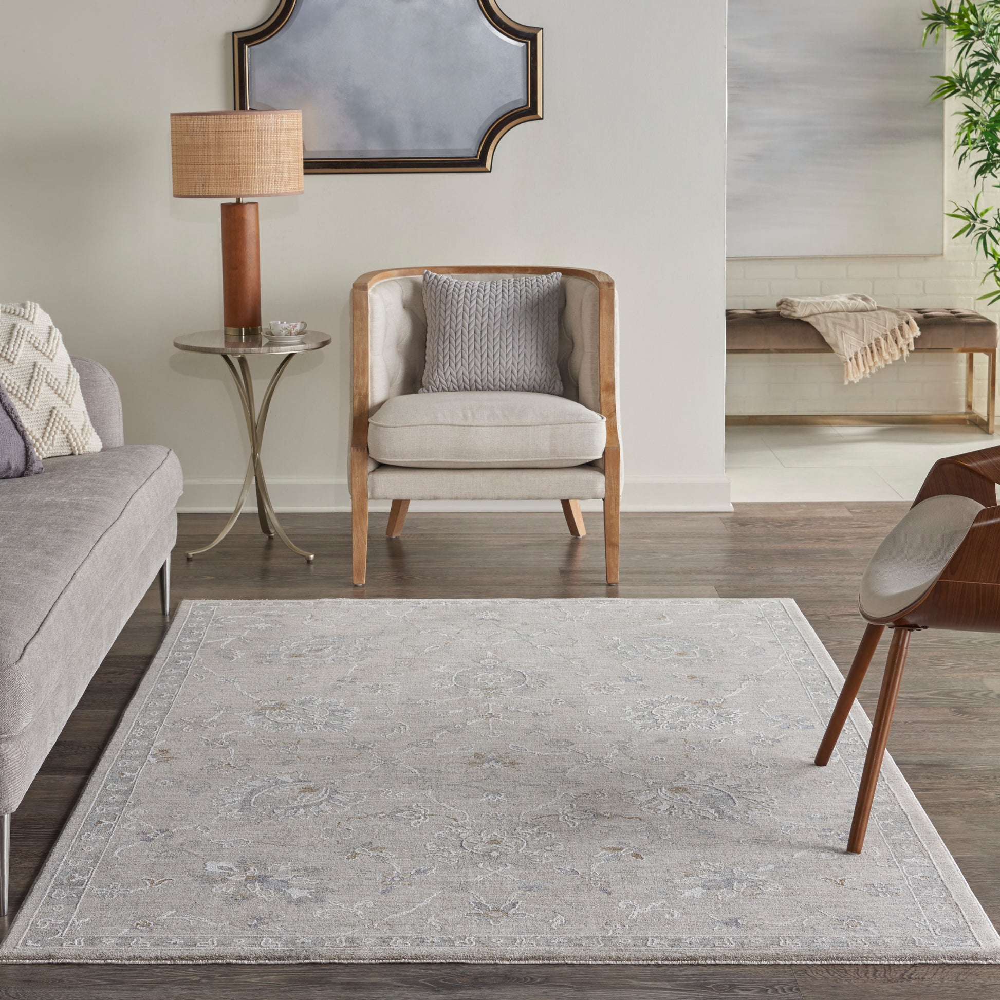 Neutral living room with gray fabric sofa, cream wood-frame chair, glass side table, wooden lamp, pale patterned rug.