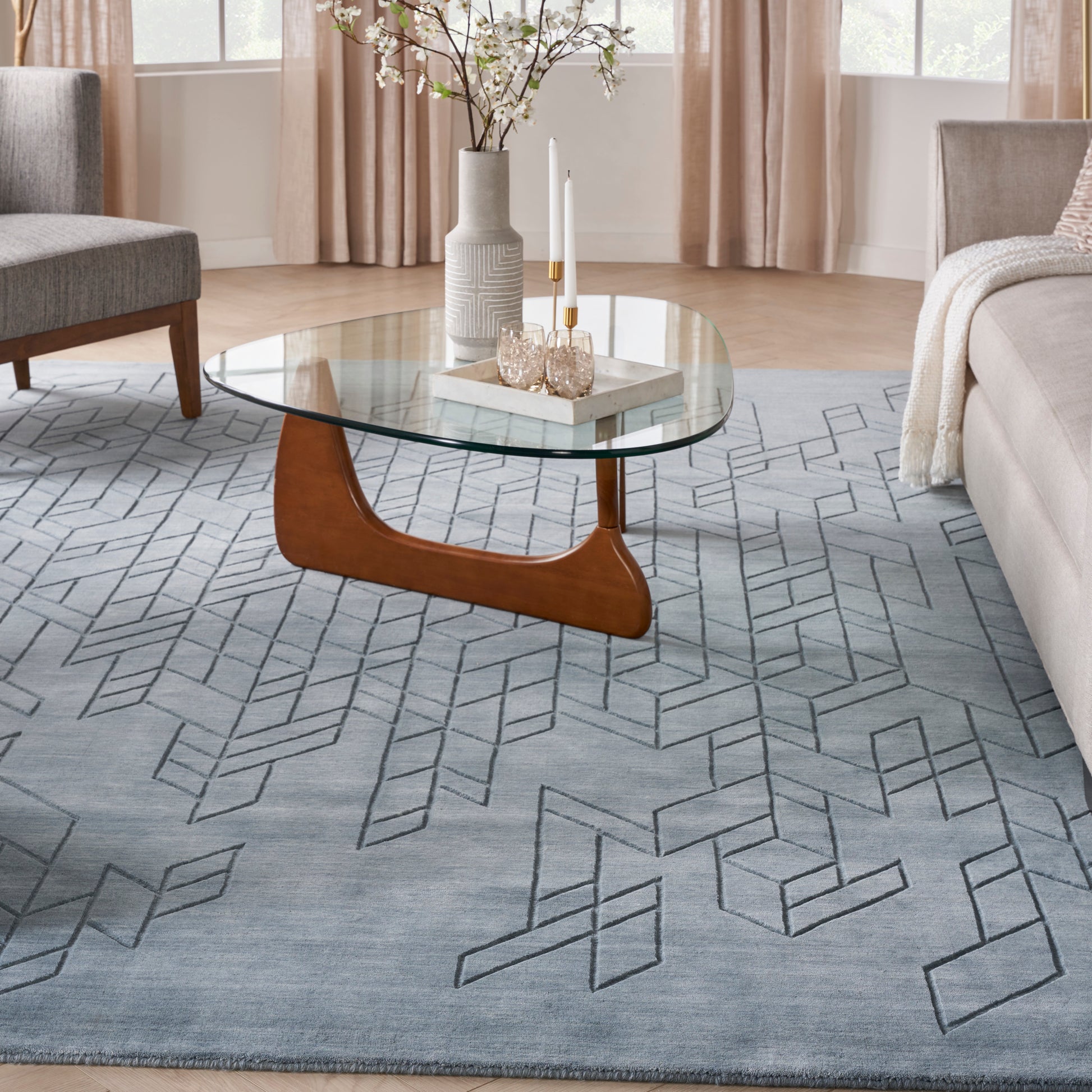Glass coffee table with curved wooden base on a geometric gray rug, vase and candles decorate the glass top.