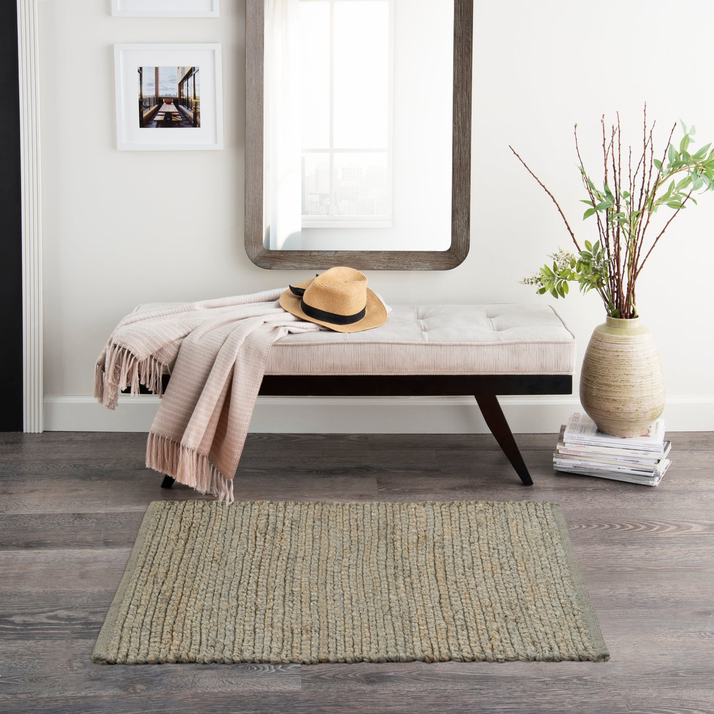 Cream tufted bench with dark wood legs, beige throw, straw hat, in front of a wooden-framed mirror; vase with branches.