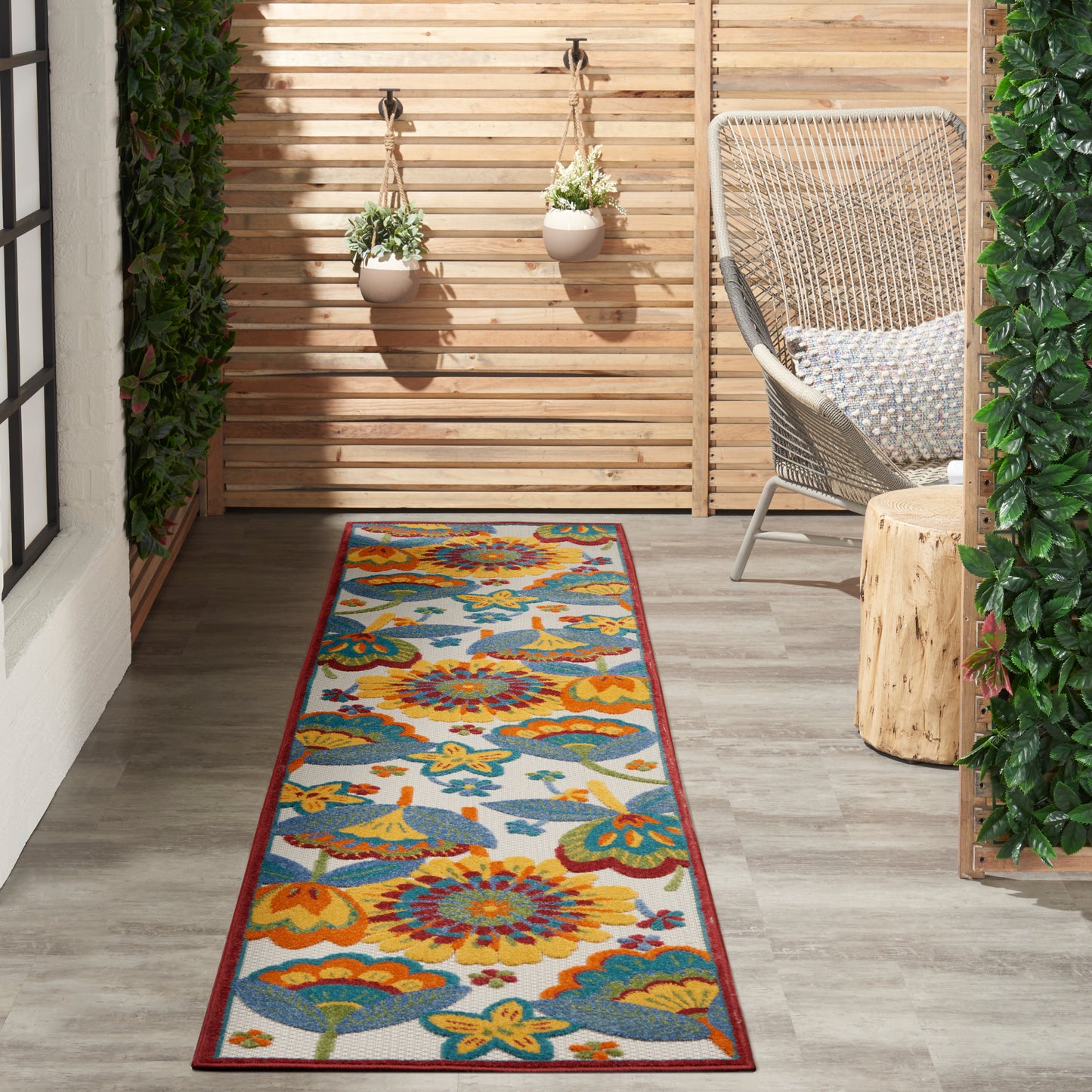 Long floral runner rug in orange, teal & yellow on a wooden deck; outdoor seating with wicker chair and hanging planters.