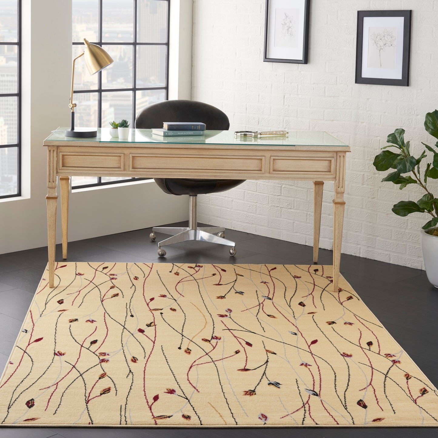 Light wood desk with glass top on a beige vine-pattern rug; black chair, brass lamp, potted plant, framed art.