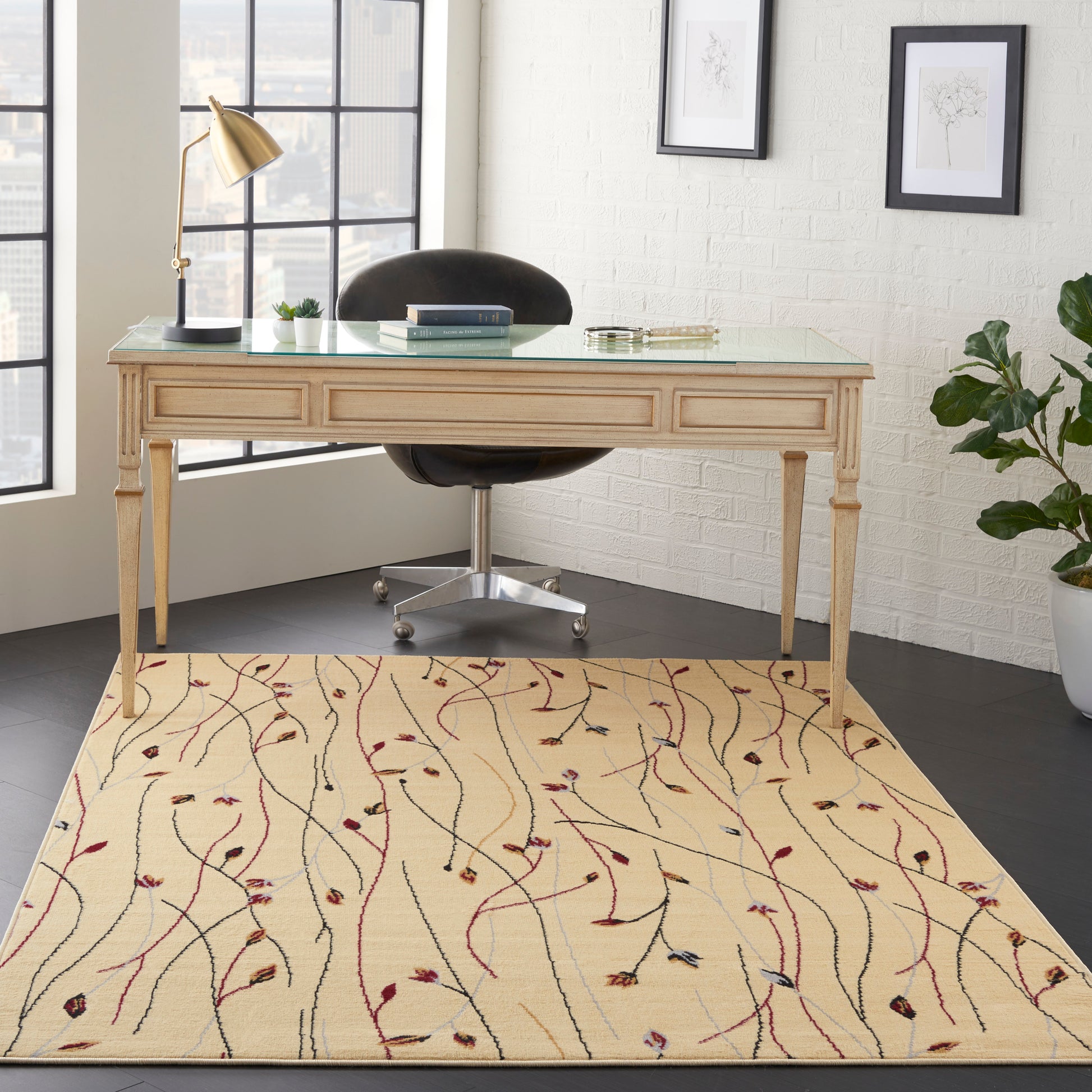 Light wood desk with glass top on a beige vine-pattern rug; black chair, brass lamp, potted plant, framed art.