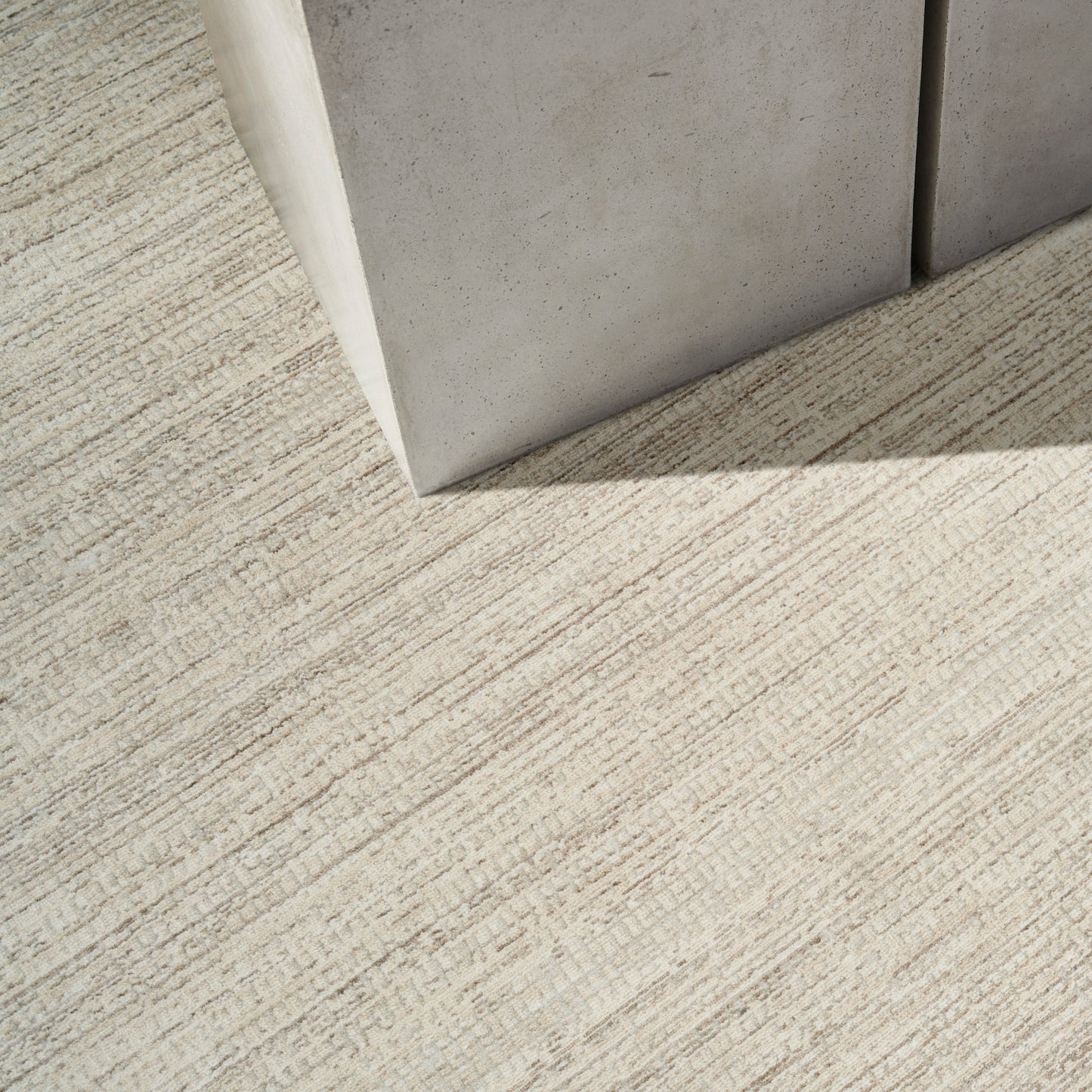 Light gray concrete corner against beige textured striped carpet with sunlit shadow.
