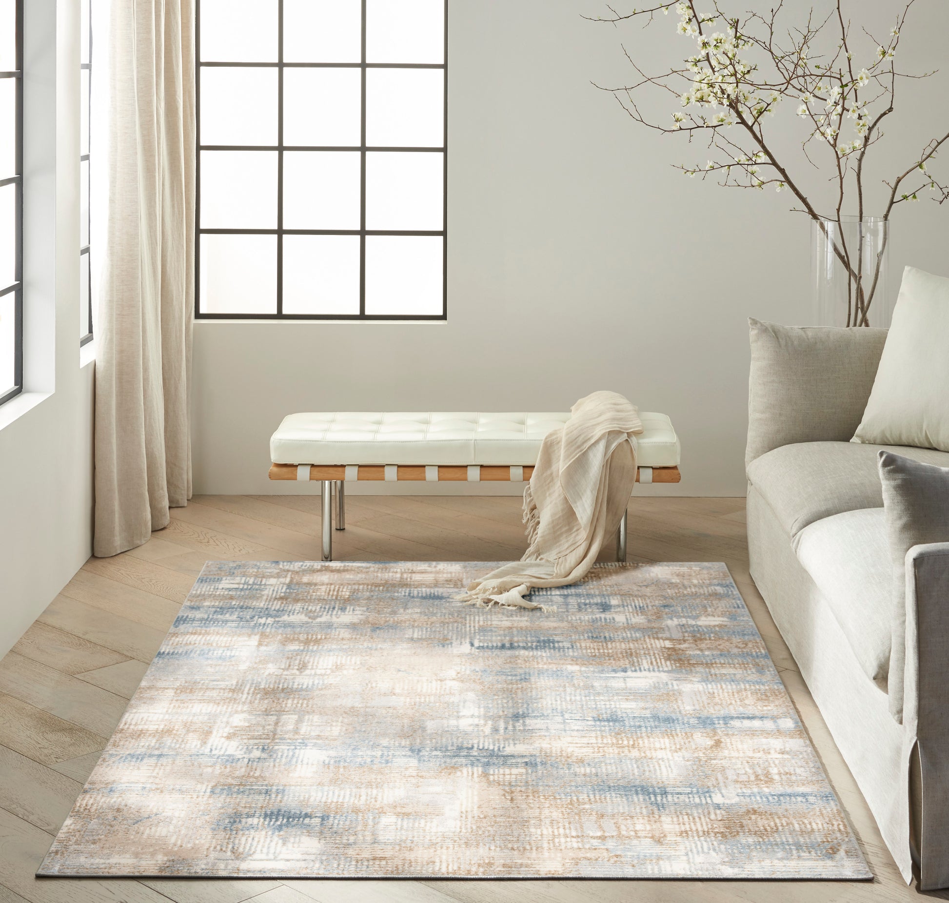 Neutral living room with a white tufted bench, beige throw, blue-gray abstract rug, light oak floor, gray sofa, grid window.