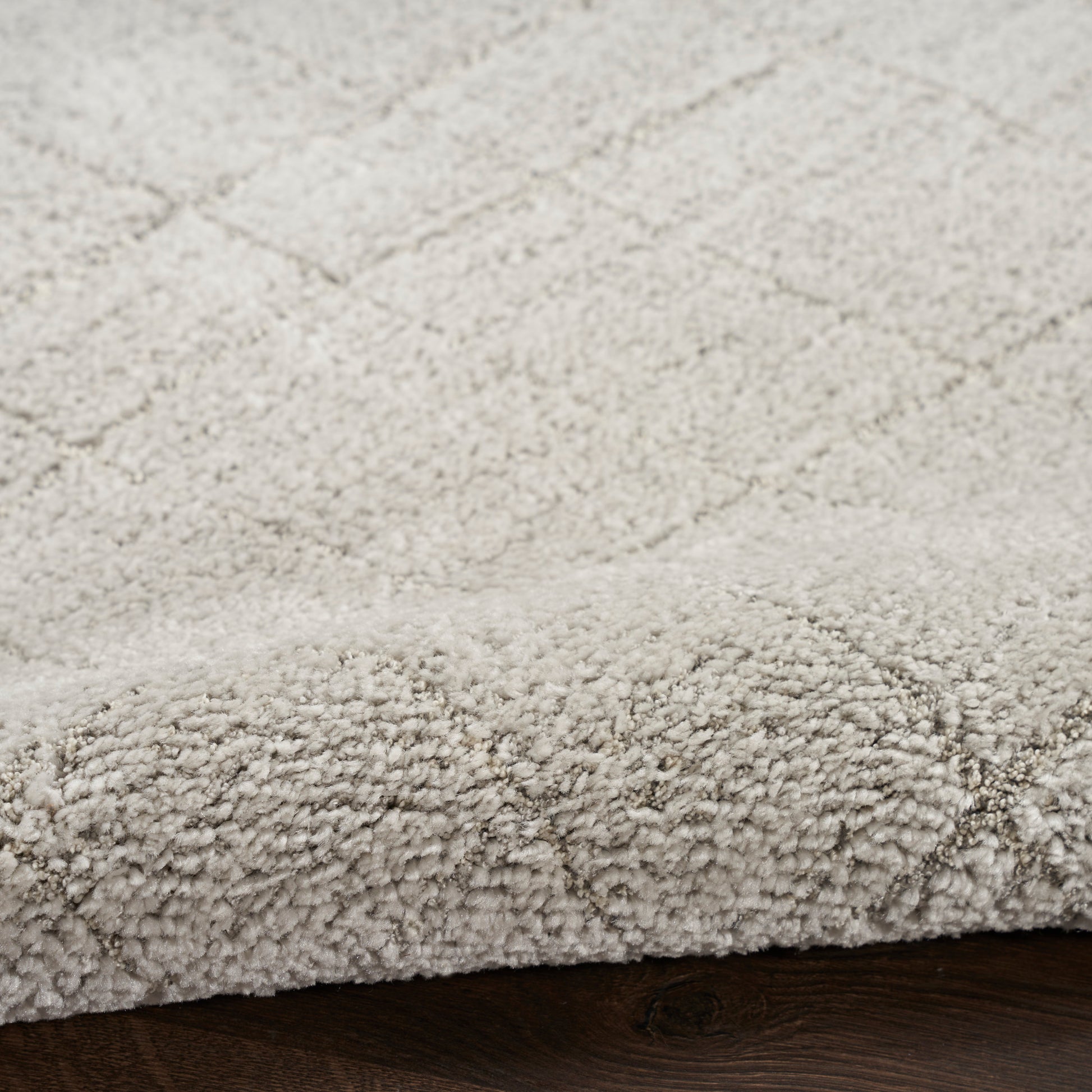 Beige plush high-pile shag rug with a subtle geometric pattern, edge on dark wood floor.