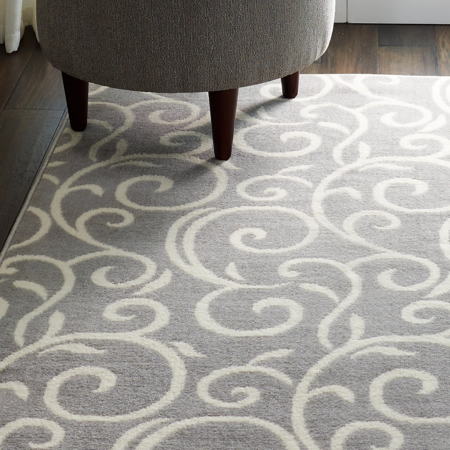 Gray area rug with white swirling scroll pattern under a gray upholstered ottoman with dark wood legs.