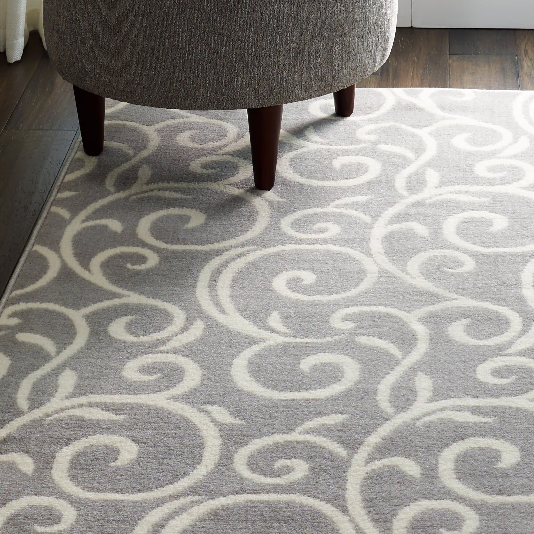 Gray area rug with white swirling scroll pattern under a gray upholstered ottoman with dark wood legs.
