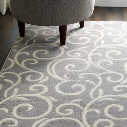 Gray area rug with white swirling scroll pattern under a gray upholstered ottoman with dark wood legs.