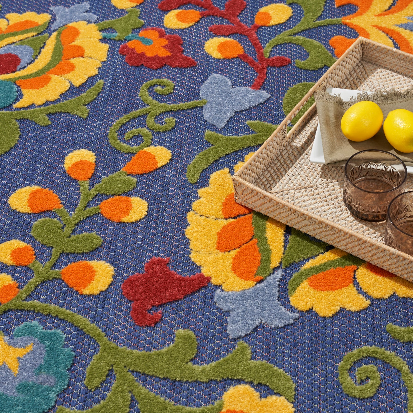Bright blue floral rug with a woven tray holding a napkin, lemons, and two amber glasses.