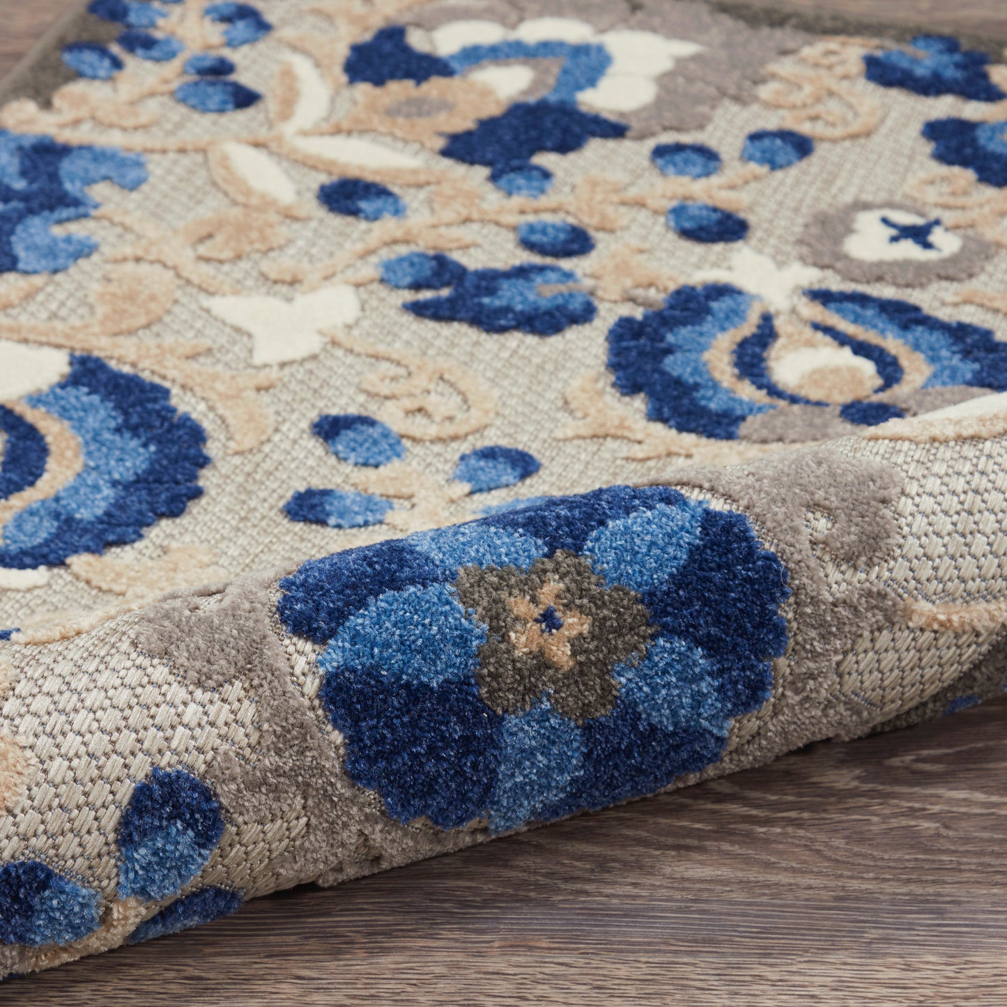 Damask-style area rug with blue flowers on beige backdrop; plush tufted pile, rolled corner on wood floor.