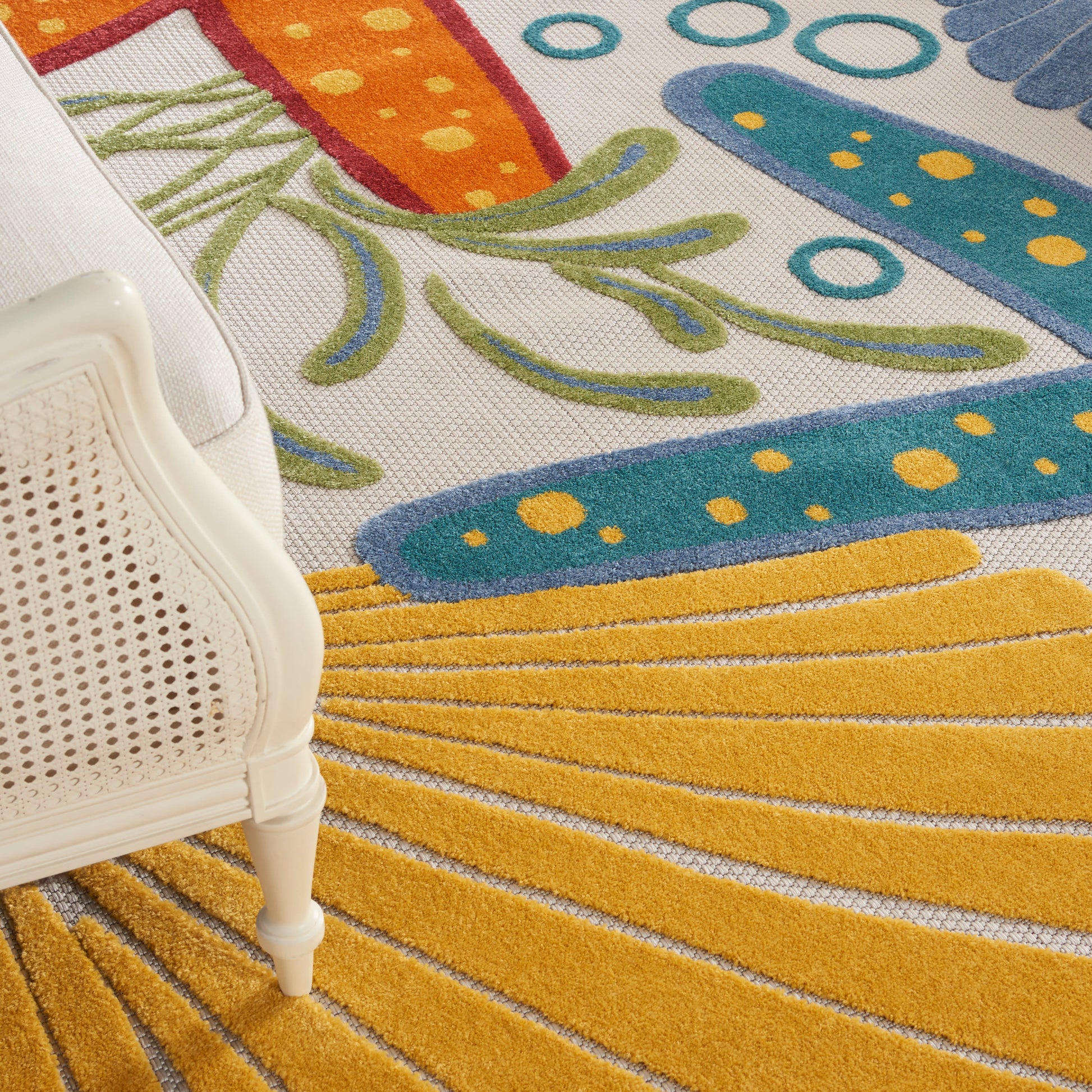 White cane-back chair leg next to a vibrant abstract floral and sunburst rug in yellow, teal, orange and blue