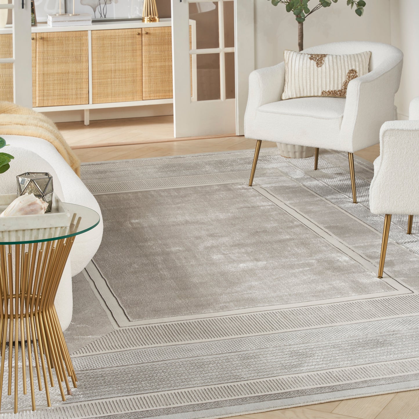 Light gray bordered area rug in a modern living room with white chairs on gold legs and a round glass-top side table.