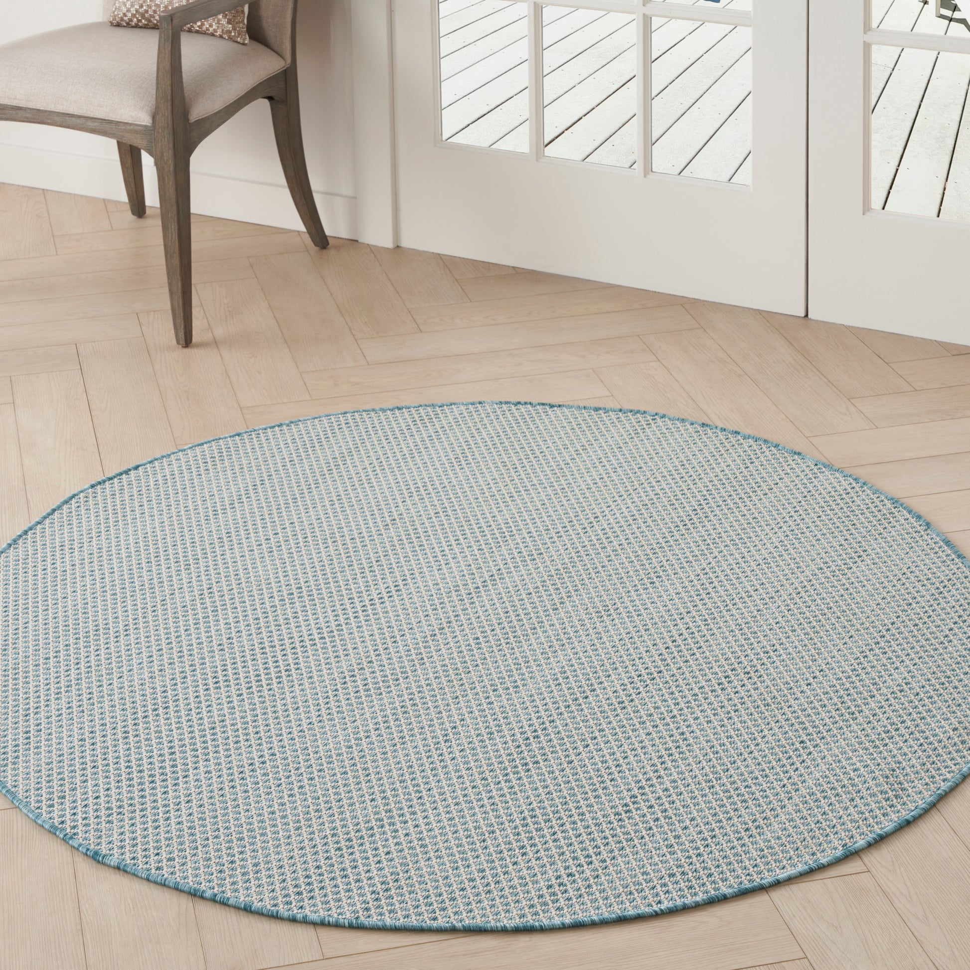 Round light-blue woven rug on pale wood herringbone floor near glass-panel doors.