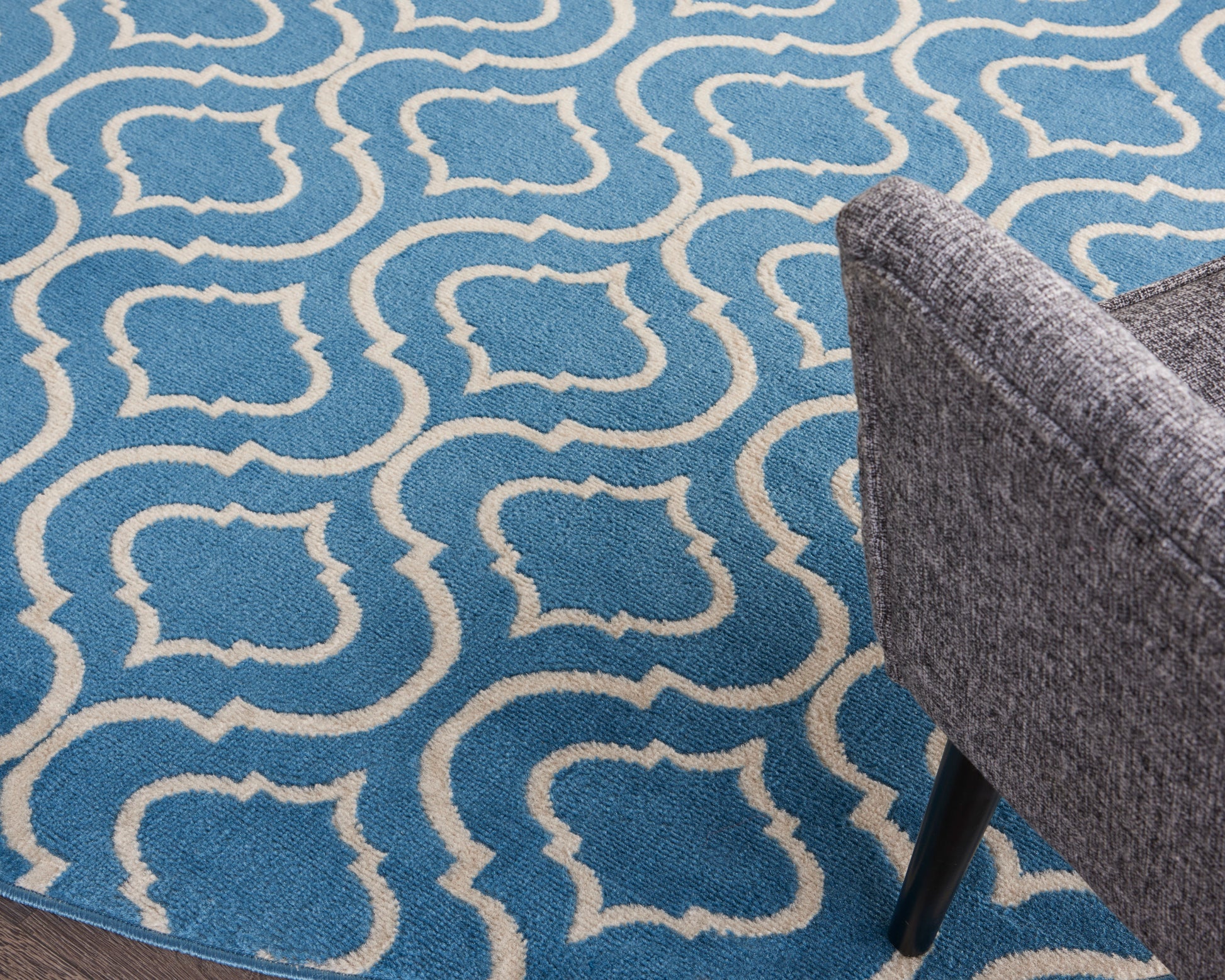 Blue rug with white interlocking geometric trellis pattern; grey upholstered chair leg visible on the right.