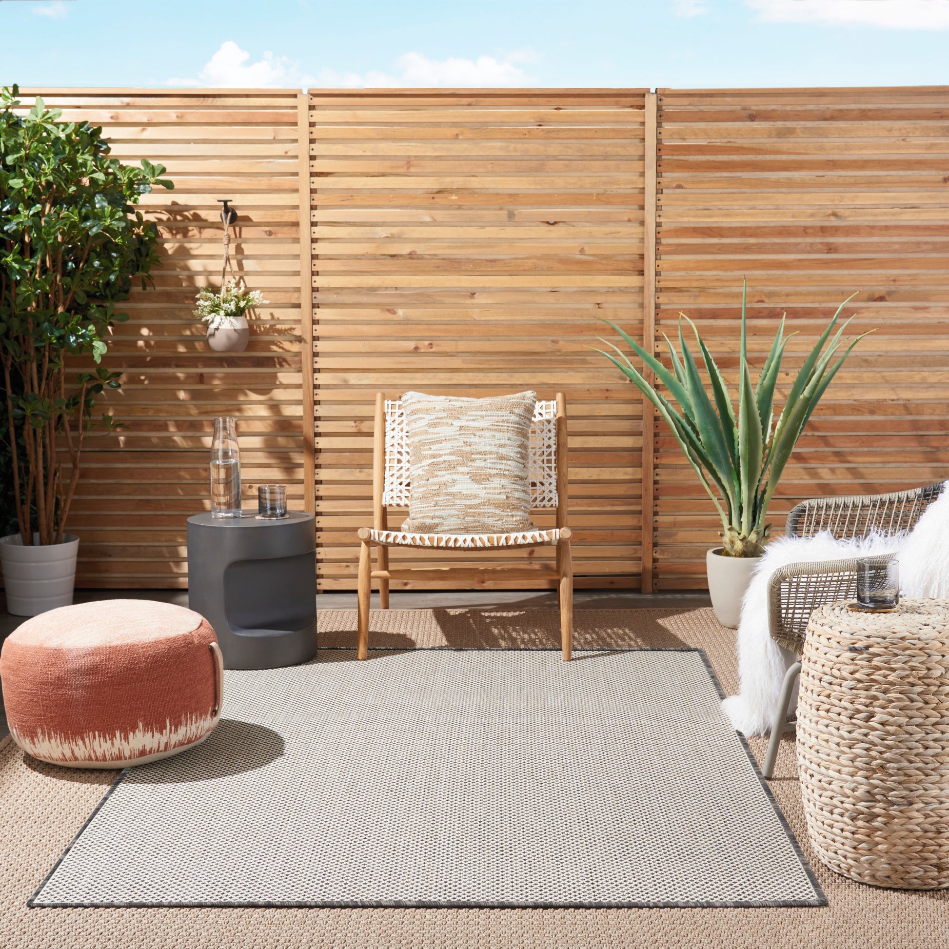 Sunny outdoor patio with wooden privacy fence, woven armchair with beige cushion, orange pouf, jute rug and tall yucca plant.