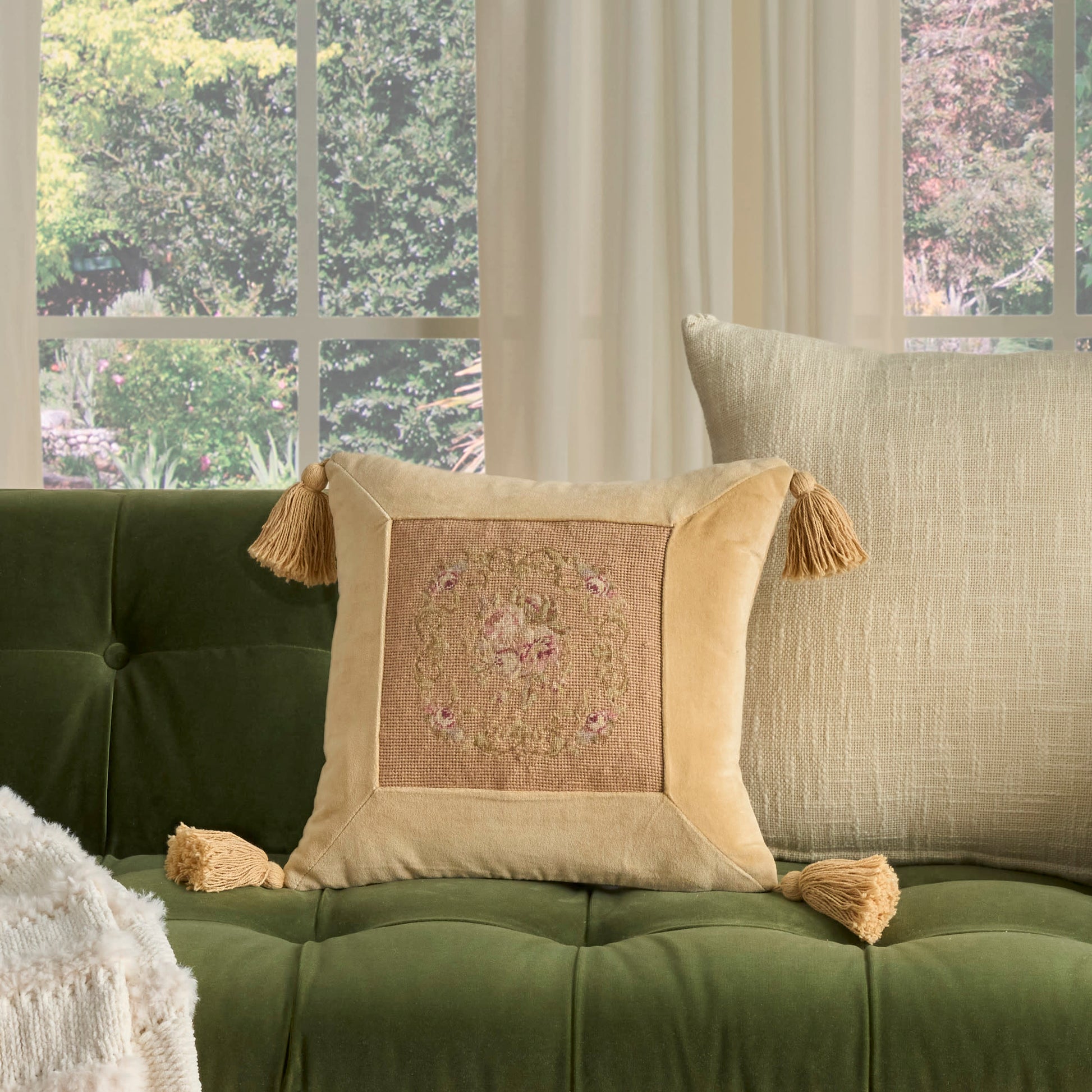 Blush decorative square throw pillow with tassels and embroidered center on a green tufted sofa in a sunlit living room