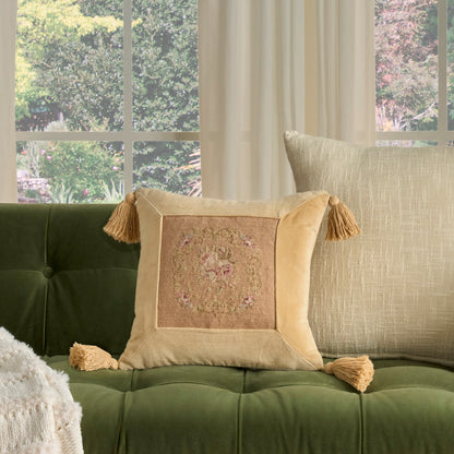 Blush decorative square throw pillow with tassels and embroidered center on a green tufted sofa in a sunlit living room