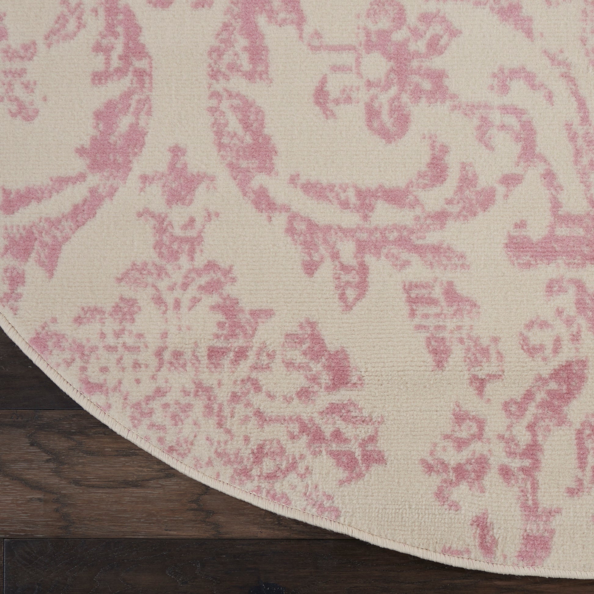 Round area rug with pink damask pattern on cream background, placed on dark wood floor.
