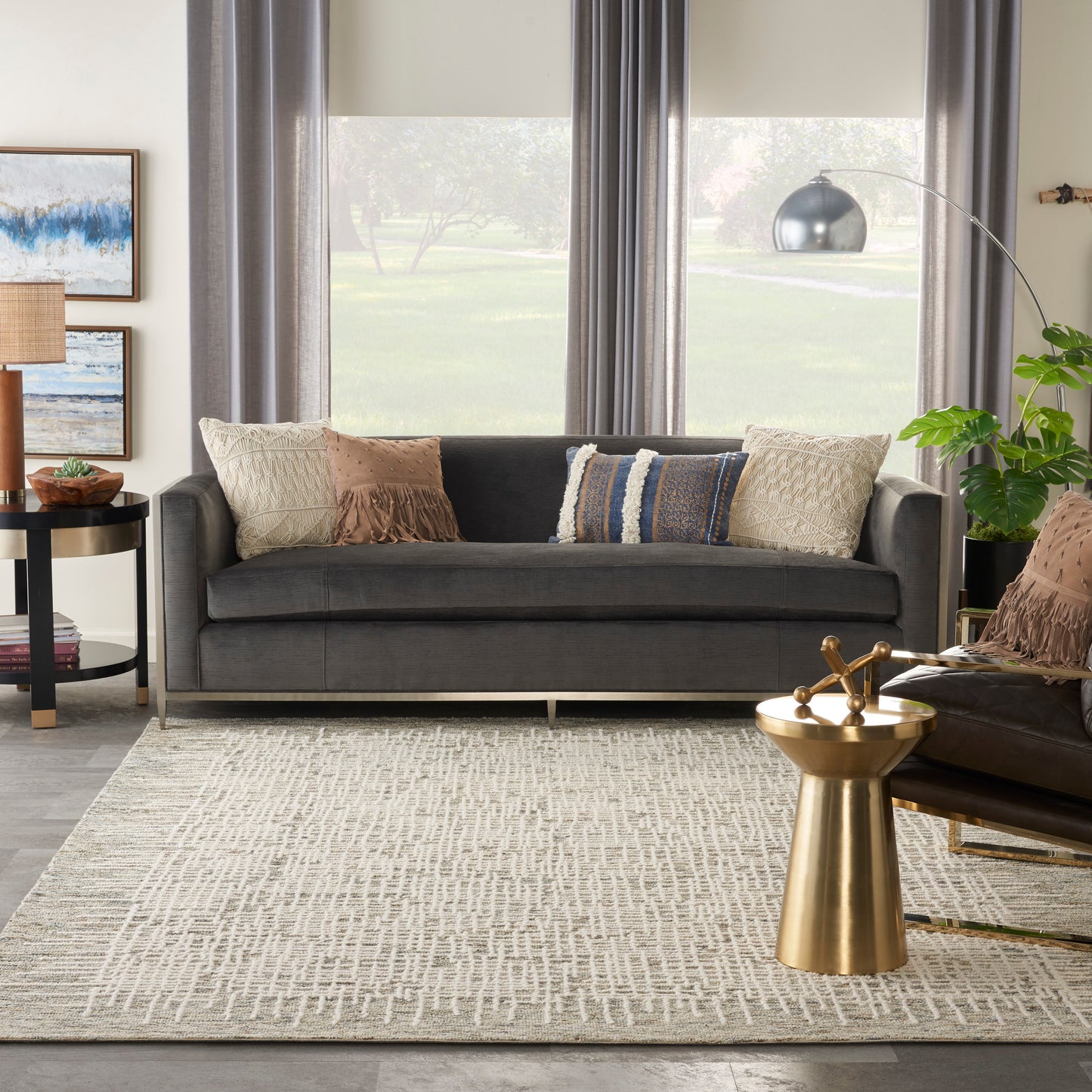 Dark gray fabric sofa with beige/tan and blue pillows, textured beige rug, gold metal side table, arc lamp, and plants.