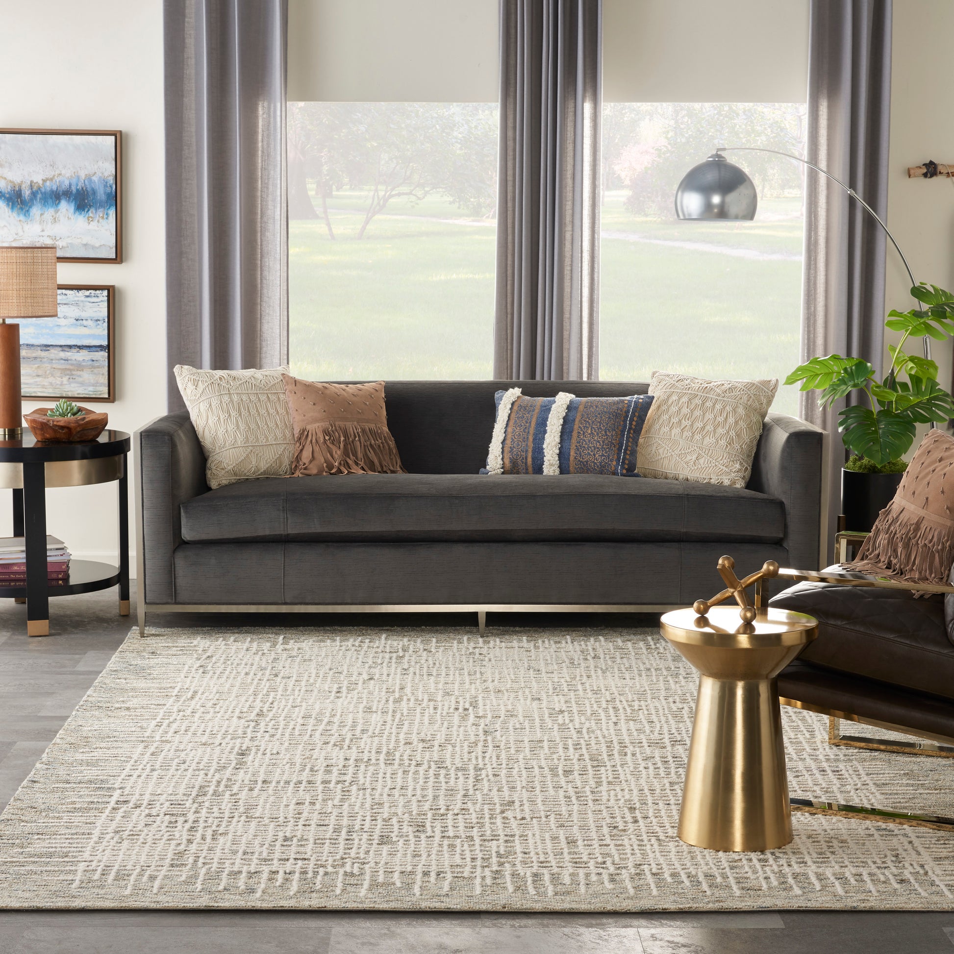 Dark gray fabric sofa with beige/tan and blue pillows, textured beige rug, gold metal side table, arc lamp, and plants.