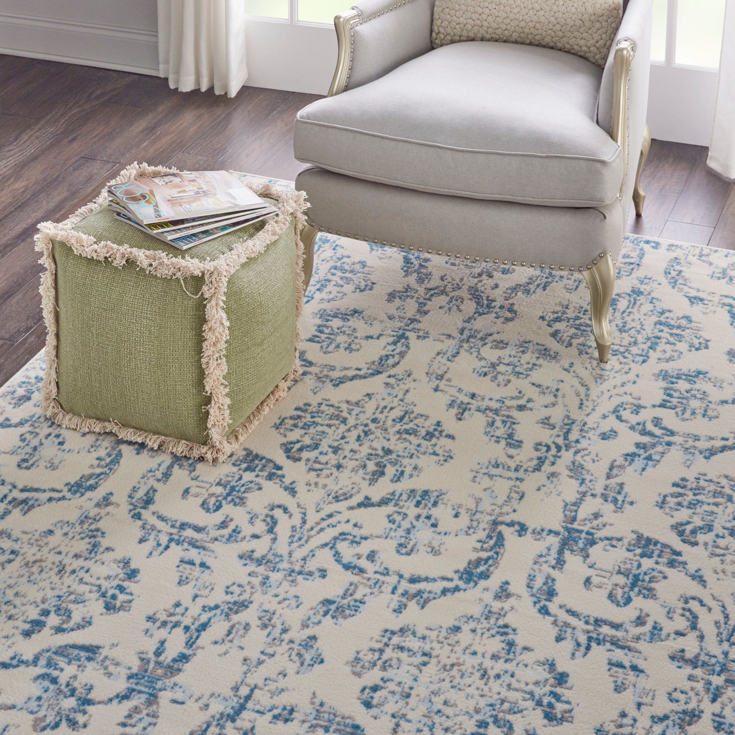 Green textured cube ottoman with cream fringe beside a light gray French-style armchair on a blue damask rug.