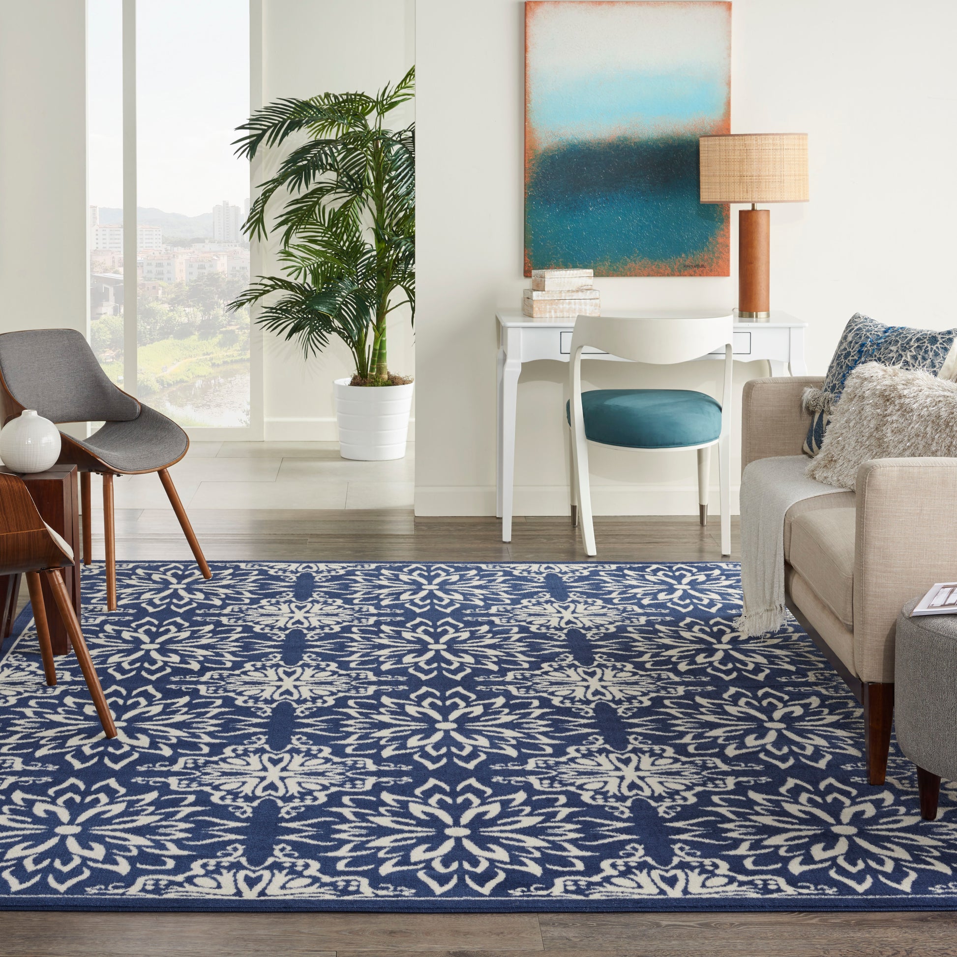 Bright living room with a blue and white patterned rug, mid-century chairs, white desk, blue chair, plant, abstract art.